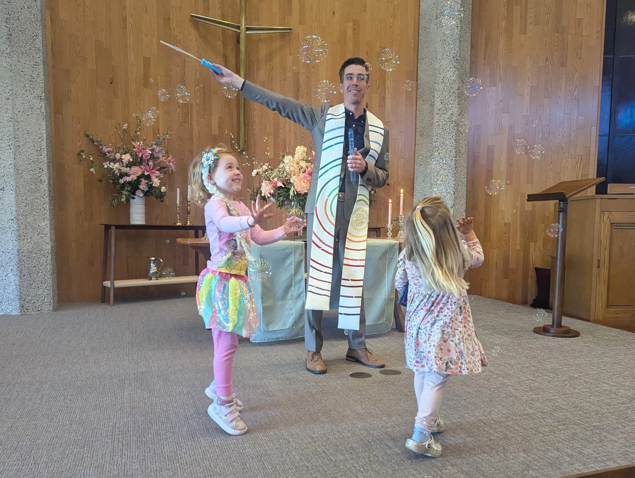 Man and two young girls inside a church, with the man holding a microphone and the girls engaging in a fun activity with bubbles, standing near floral arrangements and candles.