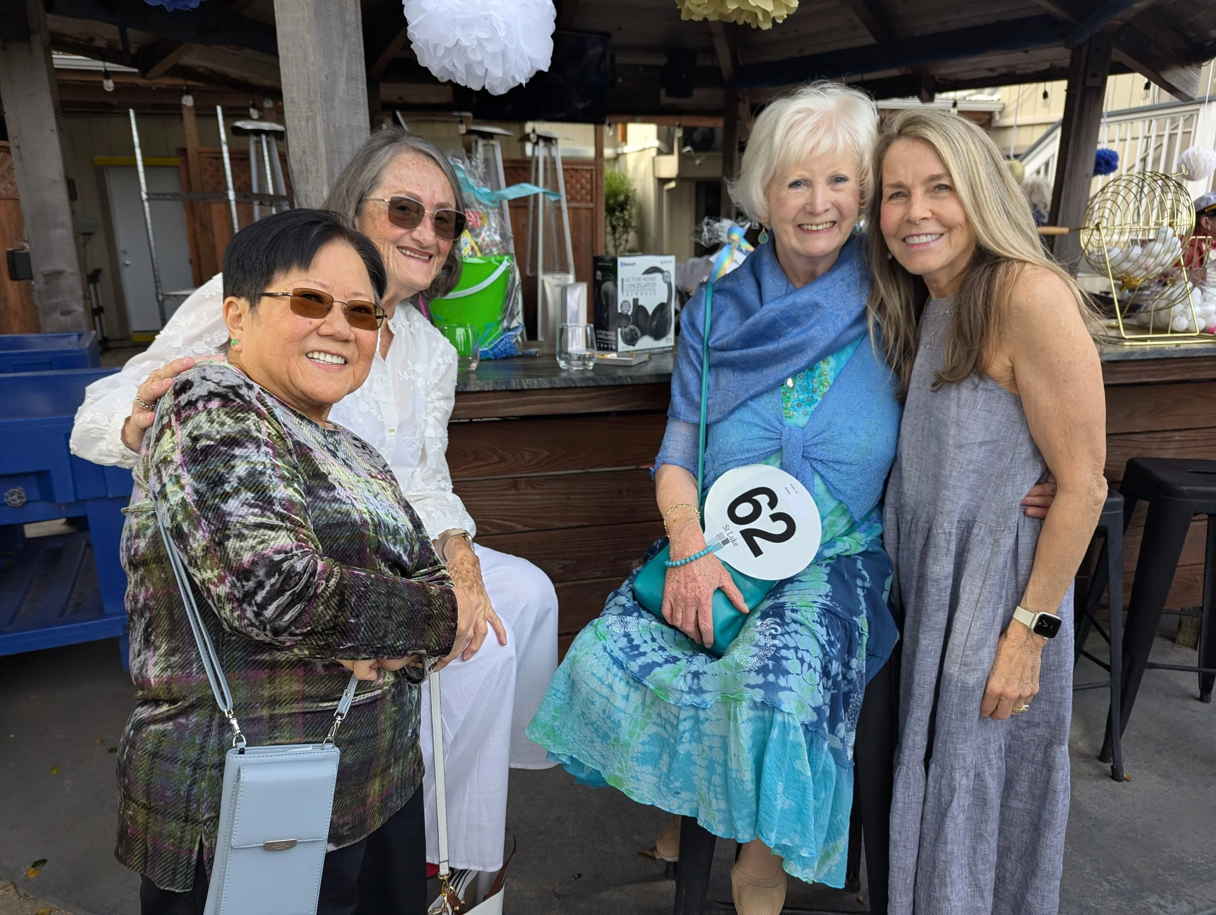 Four women posing together at an outdoor event, smiling, with decorations and a bar area in the background.