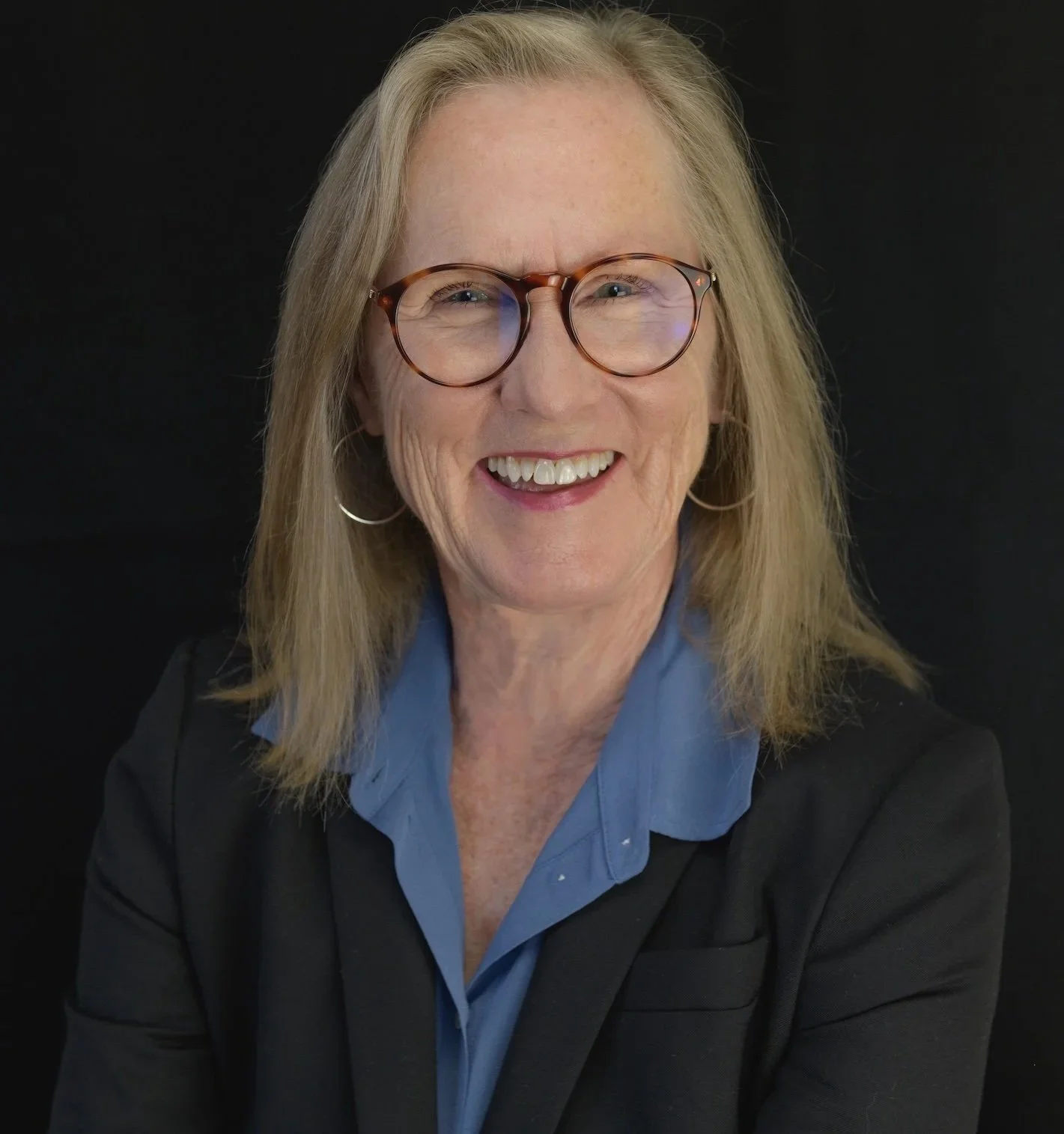 A middle-aged woman with blonde hair, glasses, and hoop earrings, smiling in front of a black background, wearing a blue shirt and black blazer.