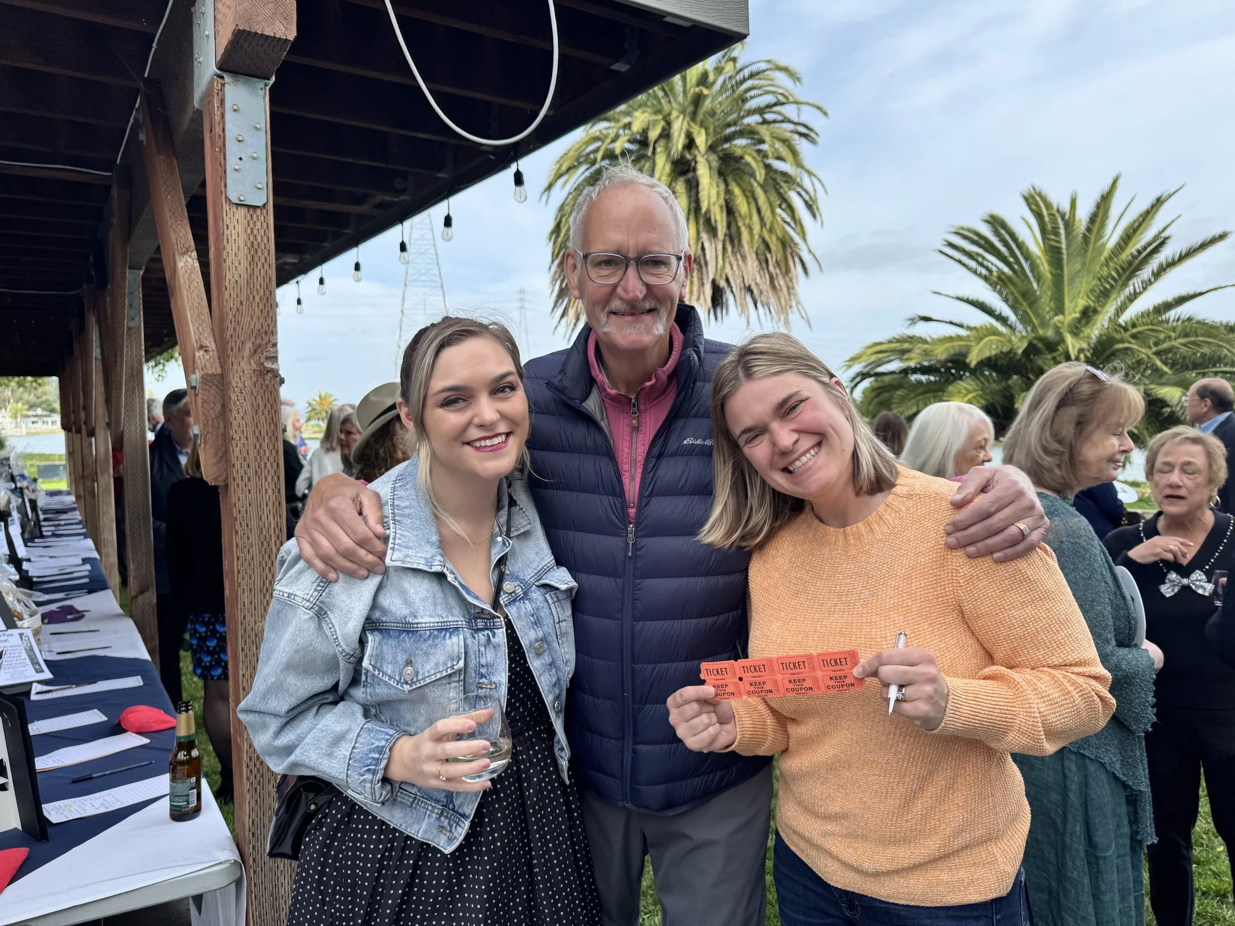 Three women and a man enjoying an outdoor event with palm trees in the background. The women are smiling, one holding a glass of wine and the other holding a group of orange tickets. The man is smiling and has his arms around the women.