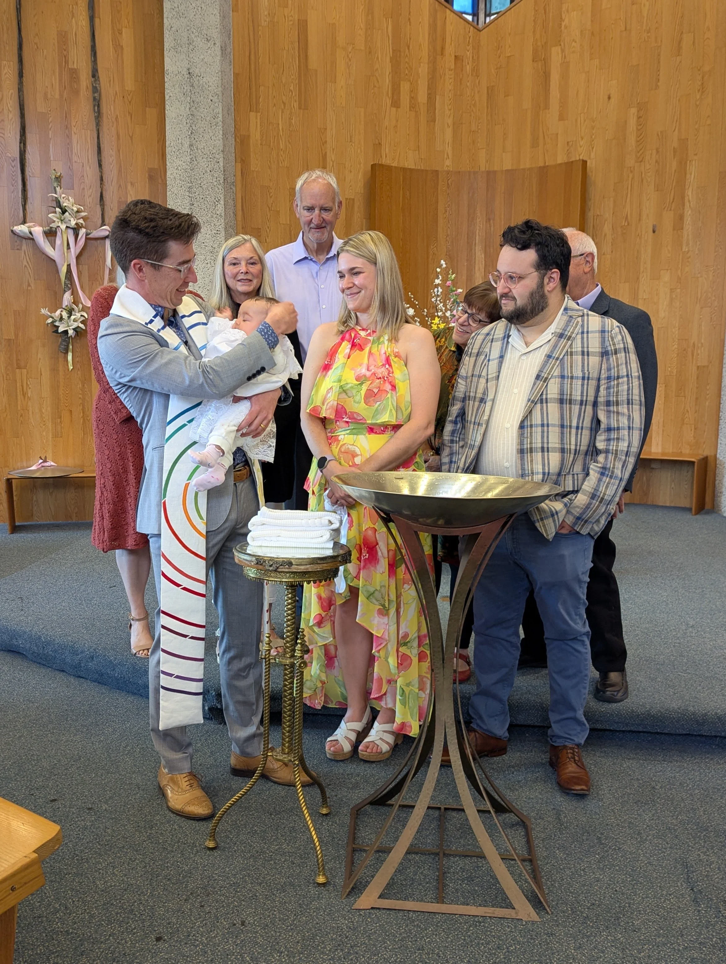 A group of people gathered around a baptismal font during a baptism ceremony in a church. The man in a gray suit is holding a baby, preparing for baptism with a woman in a colorful dress standing nearby. Several guests are watching and smiling.