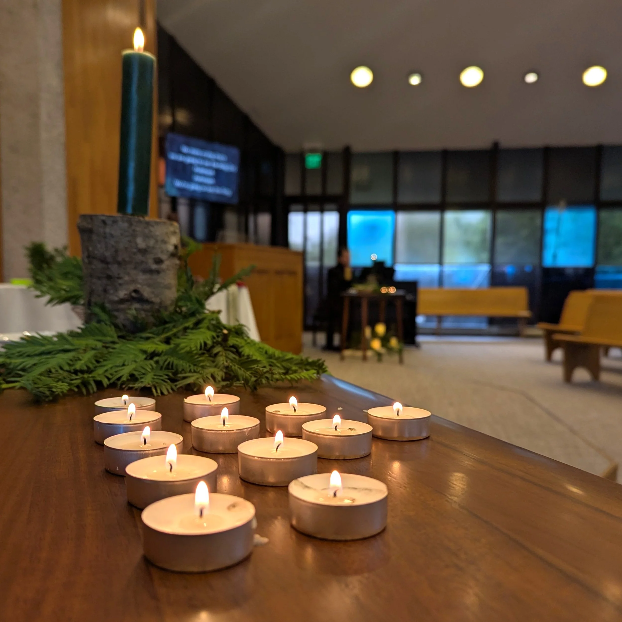 Row of small tealight candles burning on a wooden table, with greenery and candles in the background and a blurred indoor setting.