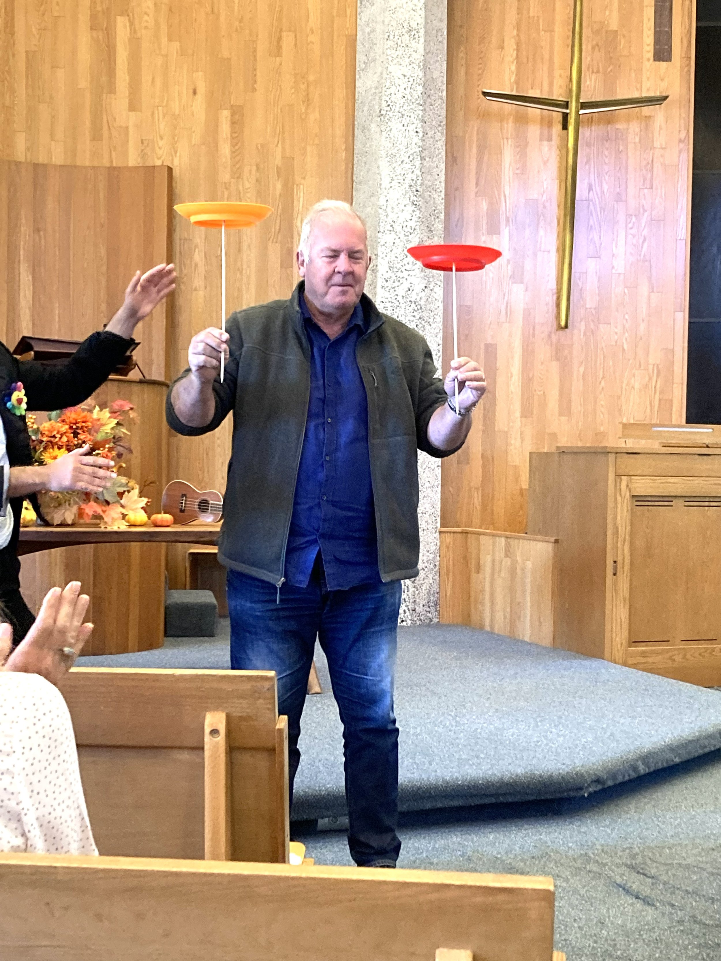 A man standing inside a church holding two dowels in each hand and spinning plates on top, one orange and one red, with a cross on the wooden wall in the background.