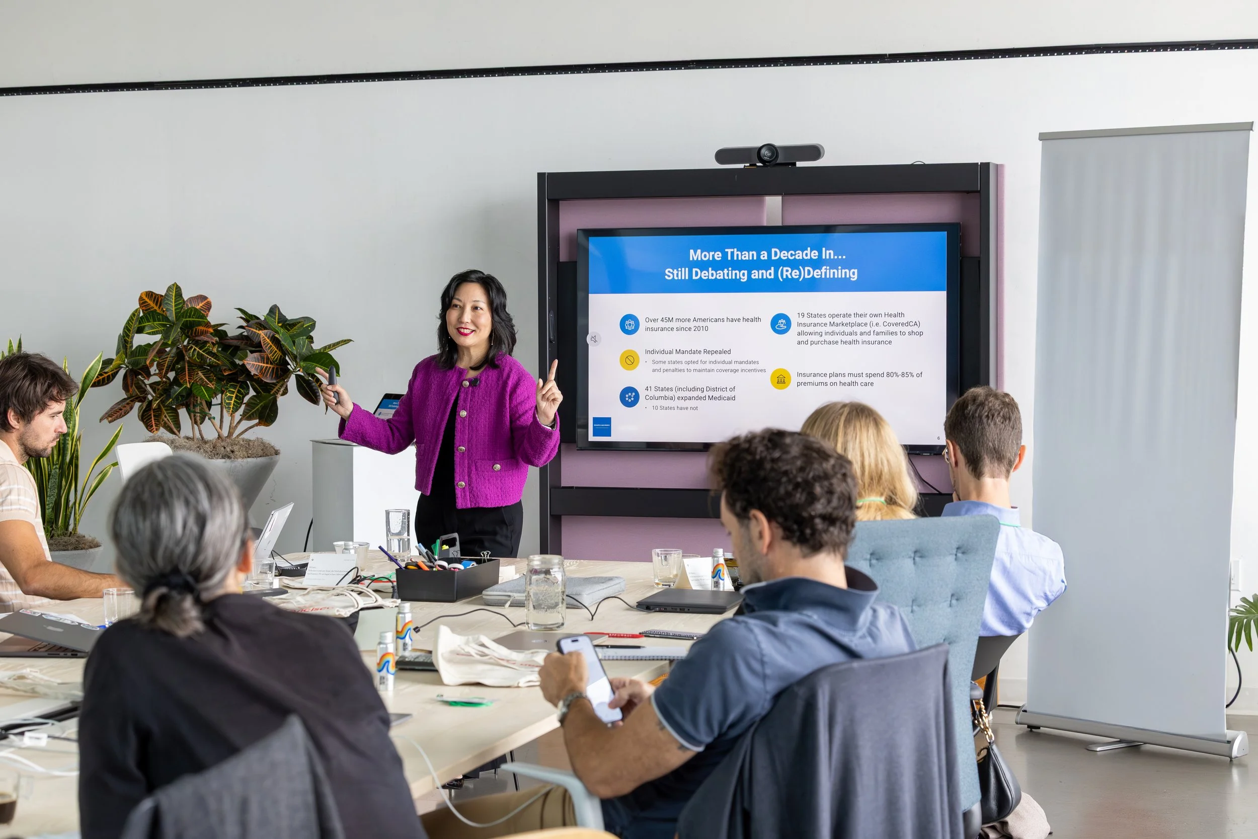 Carol Kim giving a presentation in a conference room with a large digital screen displaying health insurance data, surrounded by several seated attendees with laptops and phones.