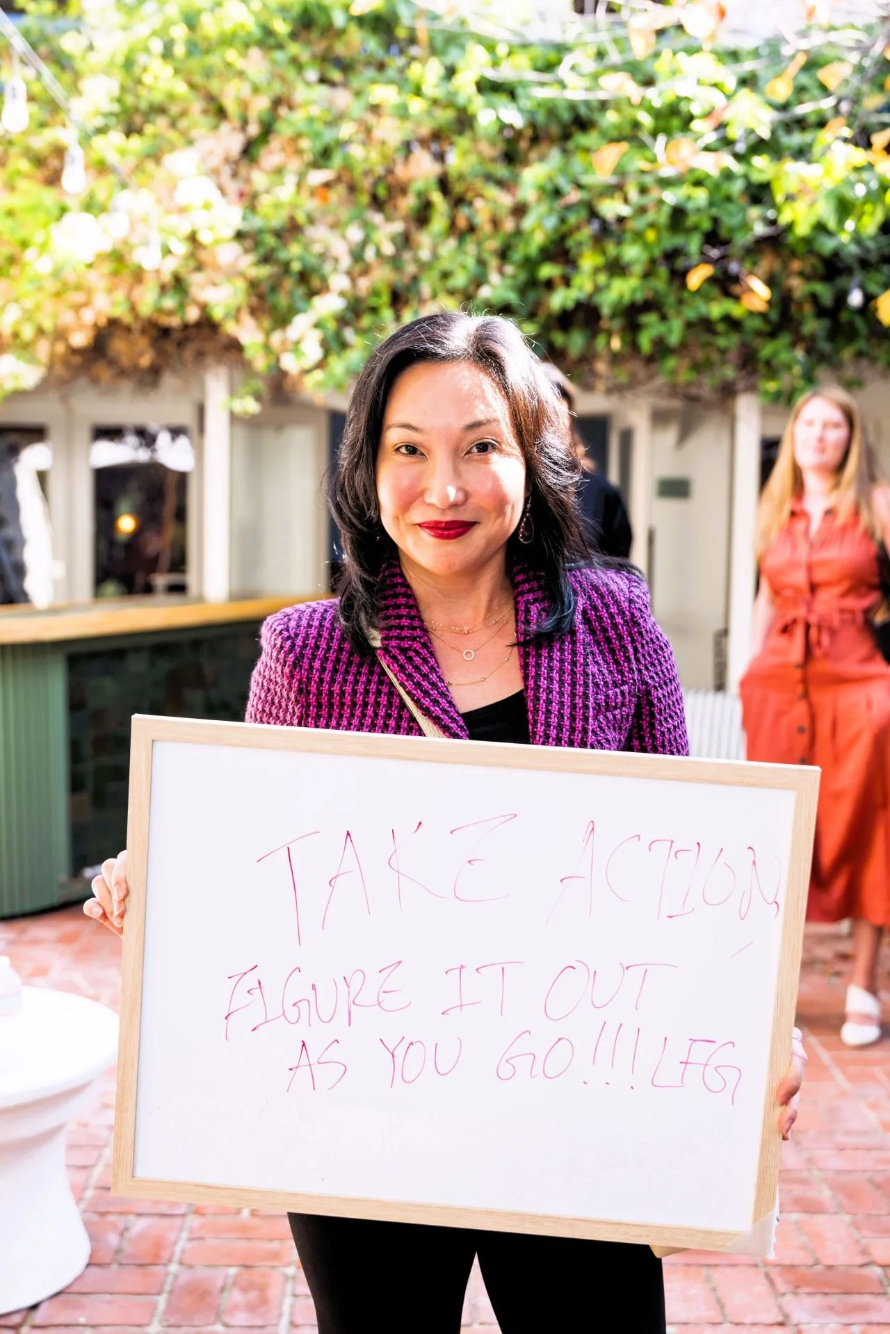 Carol Kim holding a whiteboard with the message "Take action, figure it out as you go!!!" written in pink marker, smiling at the camera at an outdoor event.