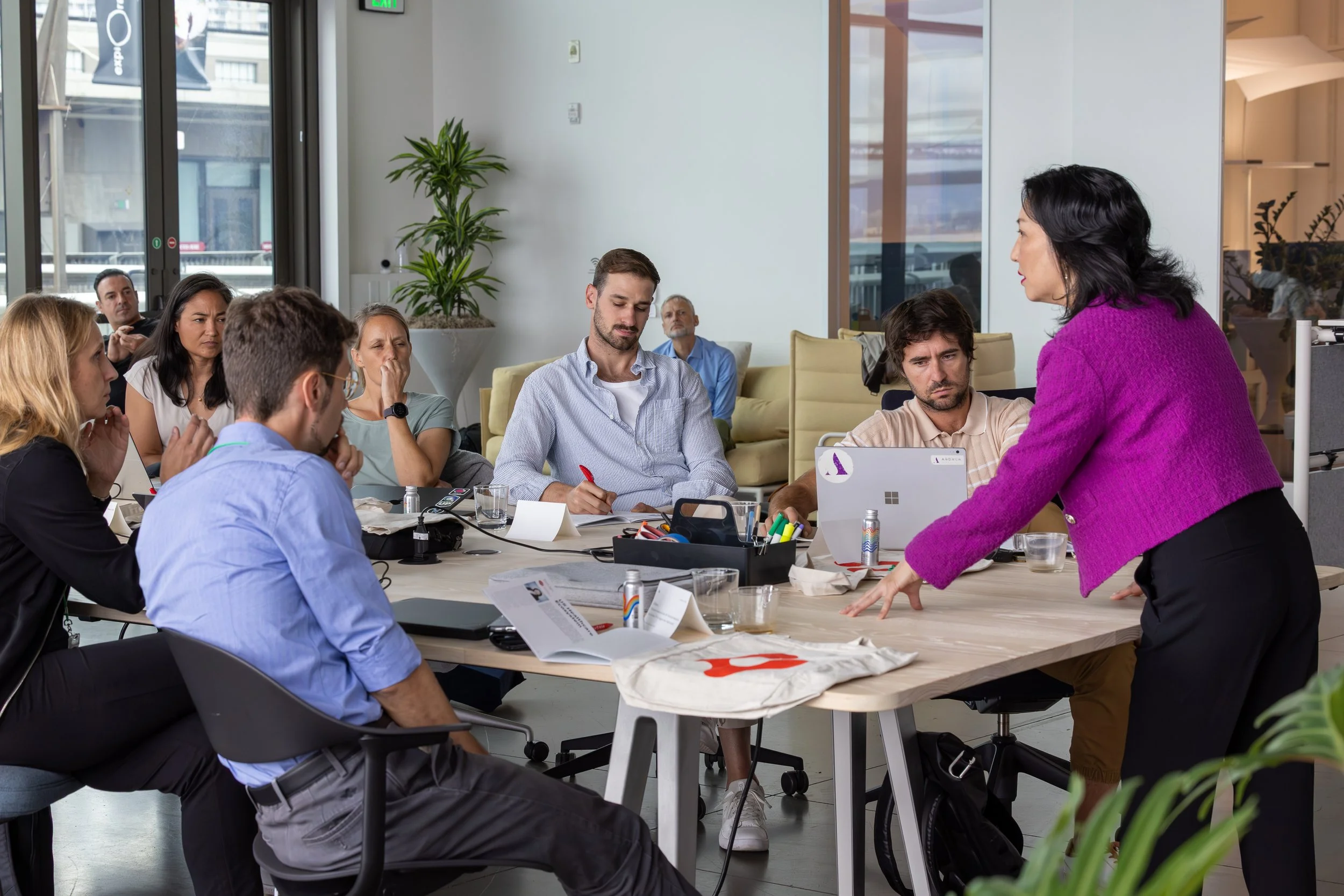 Group of people in a meeting room, listening to Carol Kim in a purple sweater who is speaking and gesturing at the table.