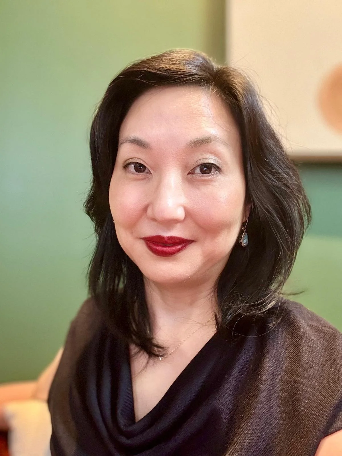 A successful entrepreneur Carol Kim with dark hair, wearing red lipstick and earrings, smiling in front of a green background.