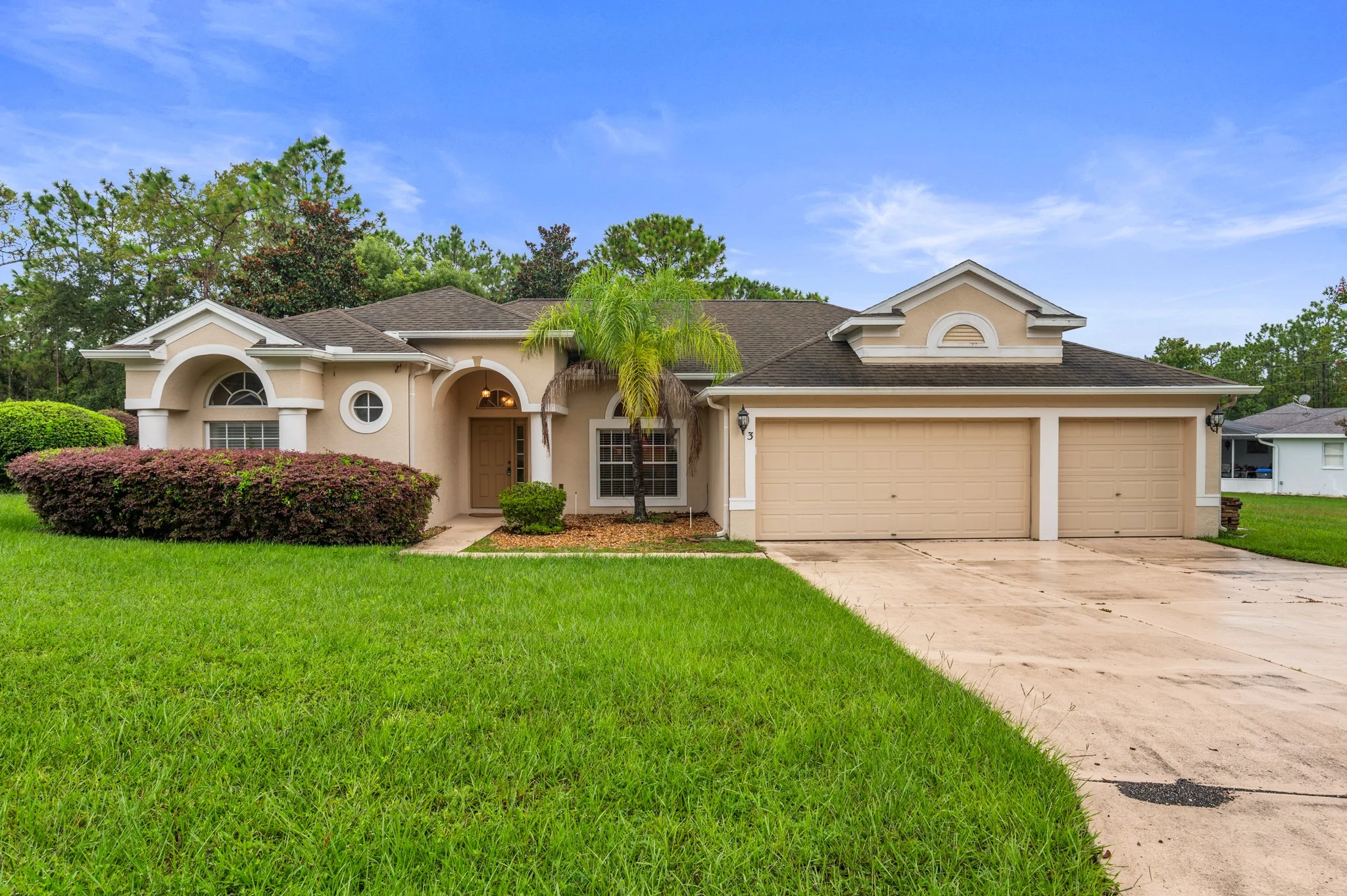 A beige stucco house with a three-car garage, green lawn, palm trees, and shrubs, under a blue sky with some clouds.