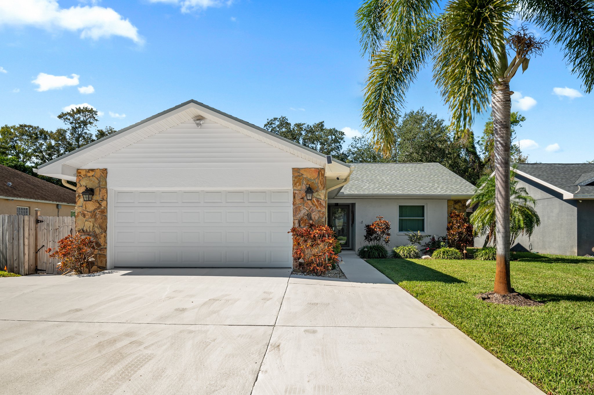 Front view of a suburban house with a white garage door, stone accents, a small front yard with green grass, palm trees, and flowering bushes, under a blue sky with a few clouds.