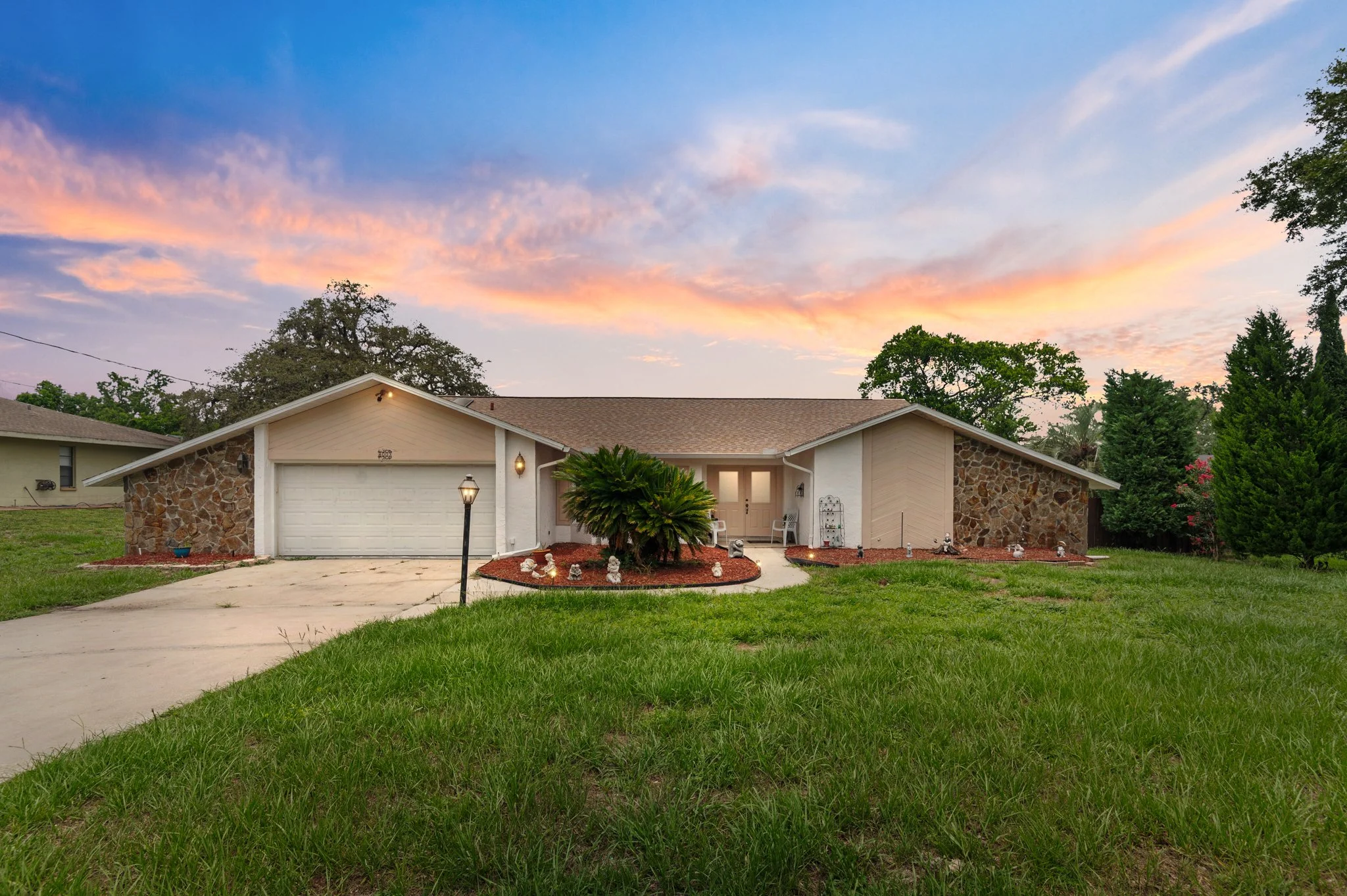 A single-story house with a stone and white exterior, front porch, and attached garage, set against a colorful sunset sky.