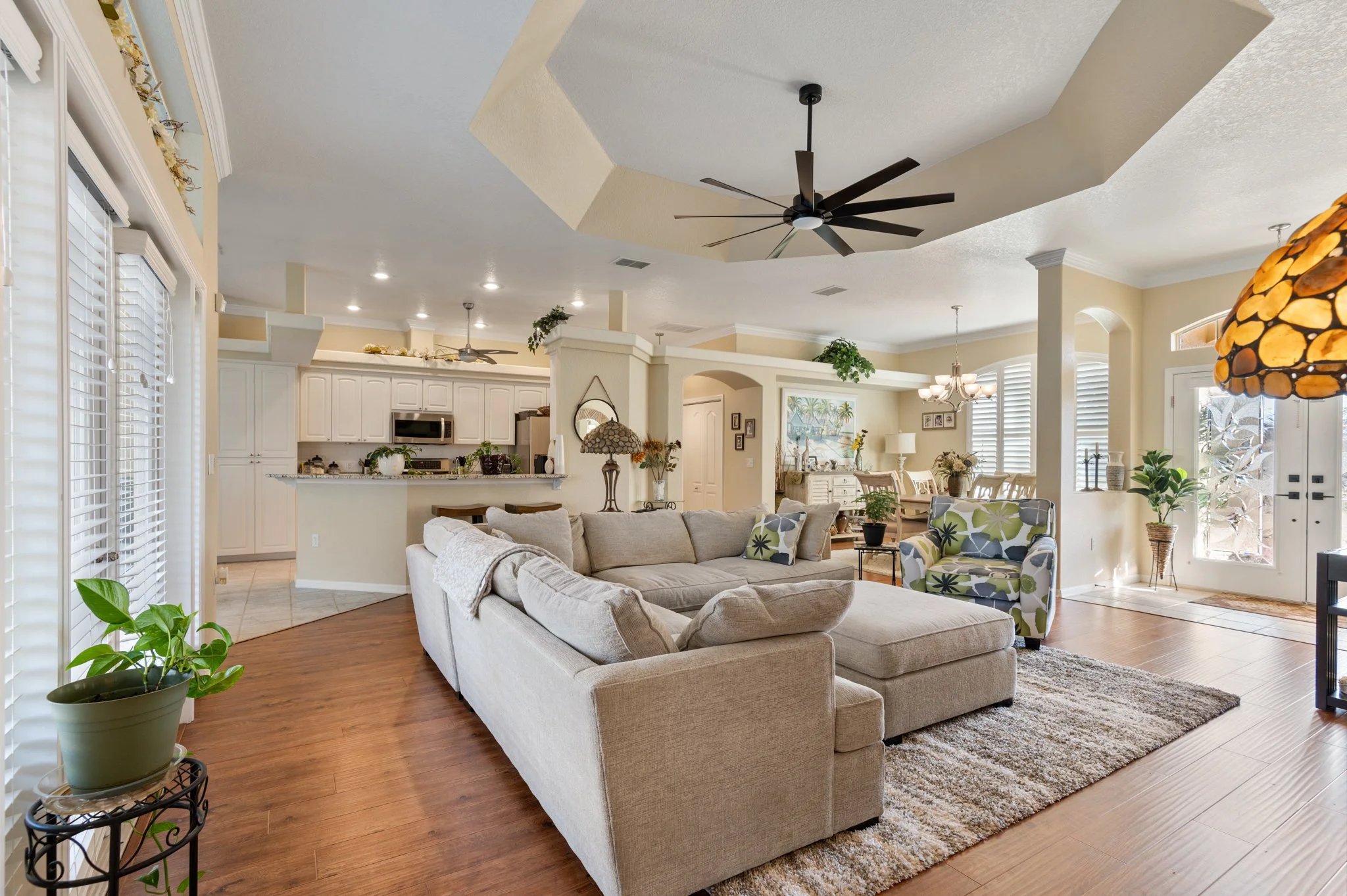 Living room with beige sectional sofa, patterned armchair, wooden floor, large windows with blinds, ceiling fan, and indoor plants.