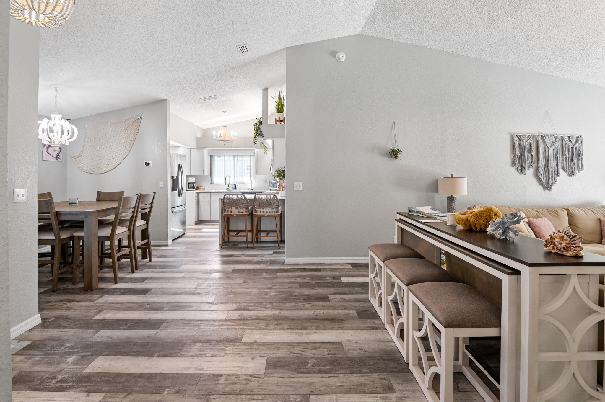 A spacious open-plan living area with a dining table, kitchen, and seating area. The dining table has six chairs, and the kitchen features white cabinetry, a window, and bar stools. The seating area includes a beige couch with decorative pillows, a s