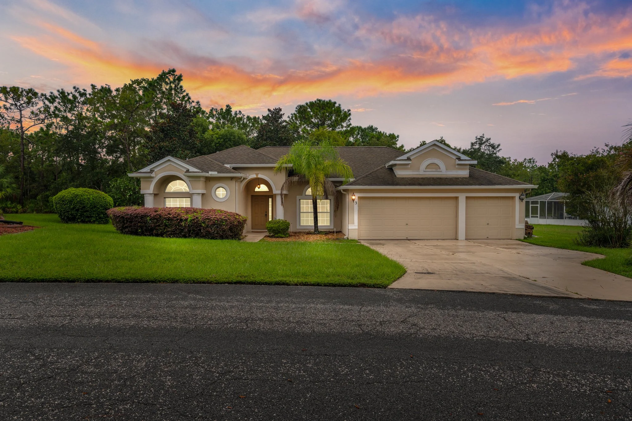 Front view of a single-story house during sunset, with a well-maintained lawn, bushes, and trees, and a driveway leading to a two-car garage.