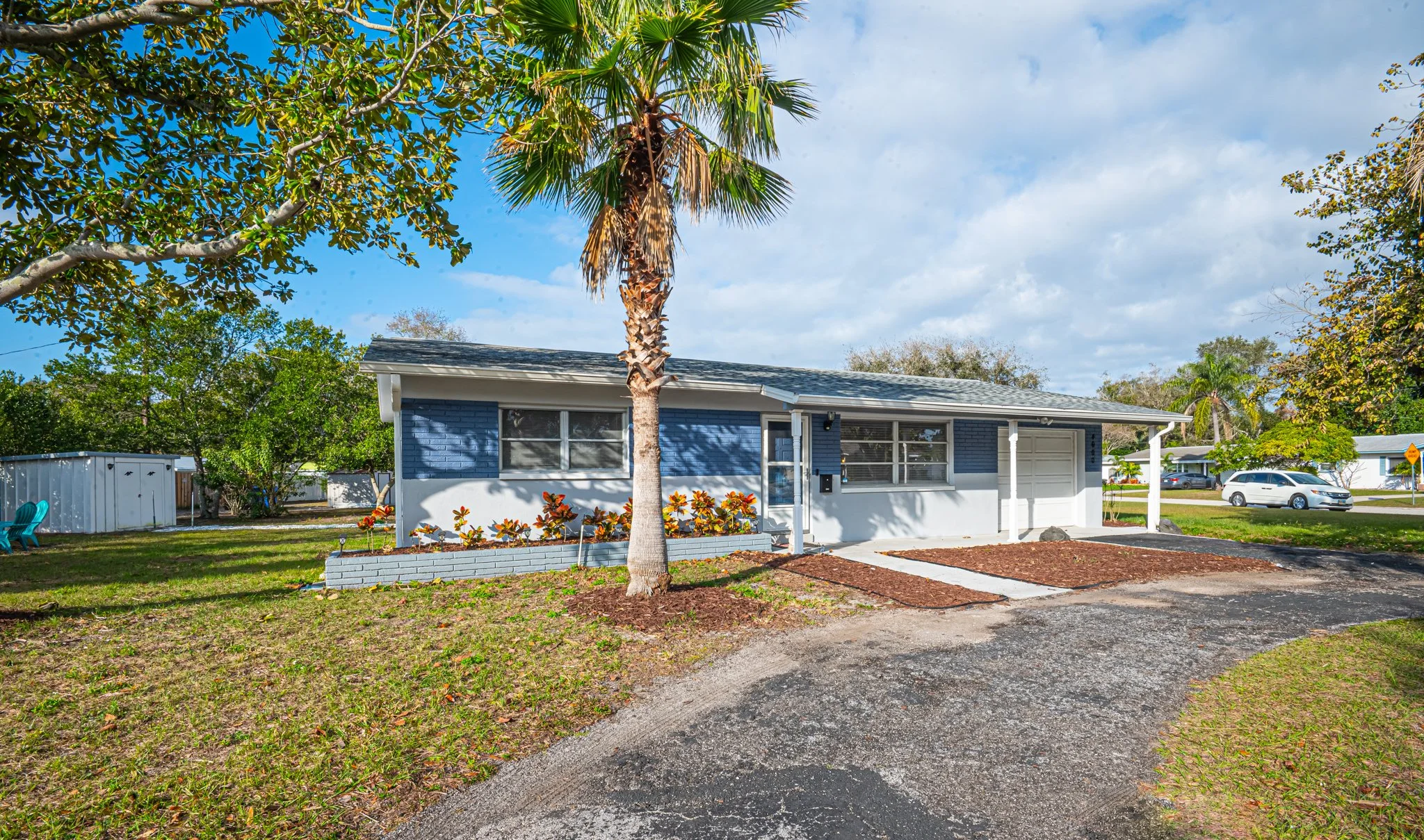 Front view of a single-story house with blue and white exterior, a driveway, a tall palm tree, and landscaping under a partly cloudy sky.