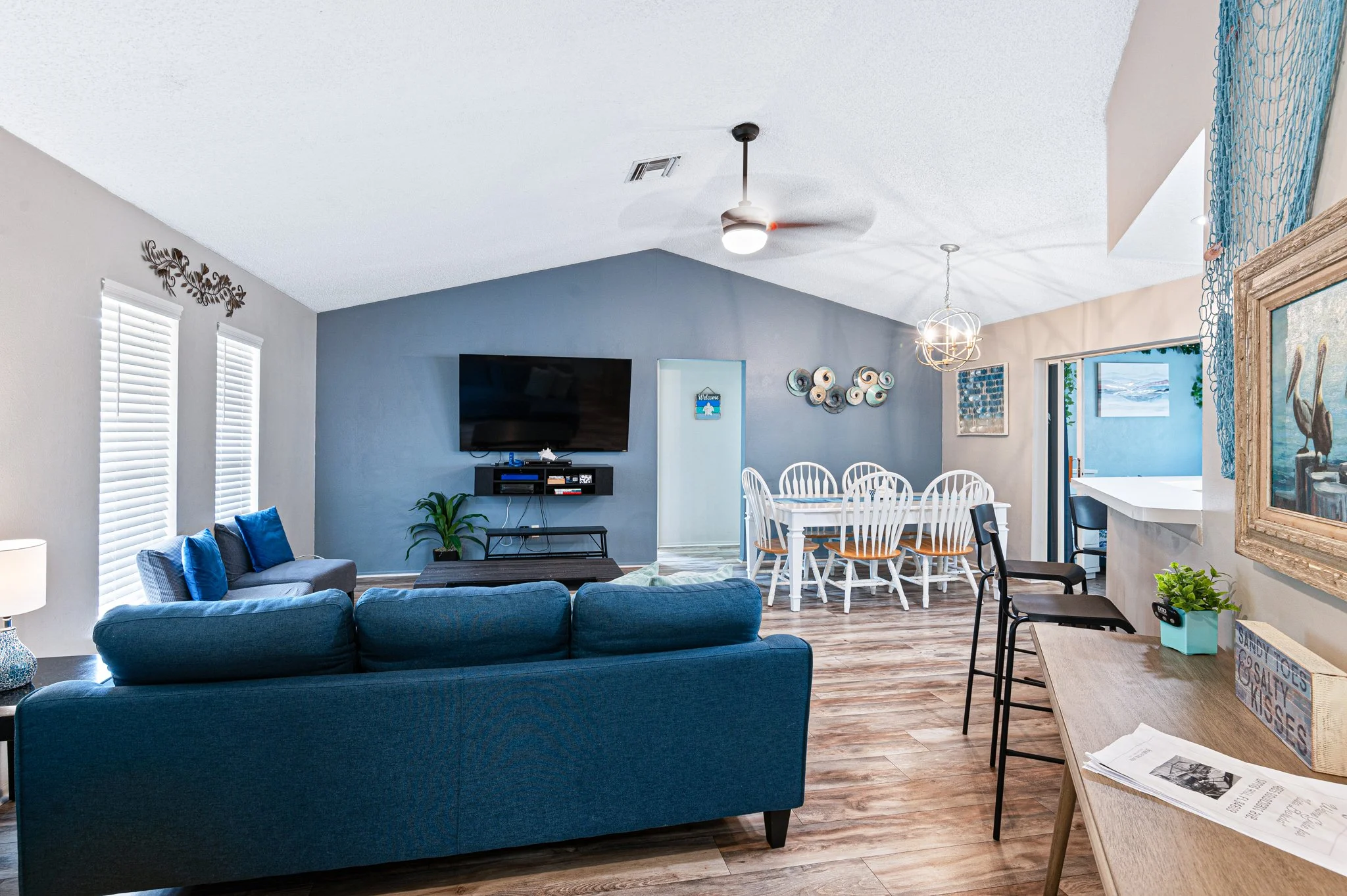 Open-concept living and dining area with blue sofa, wall-mounted TV, dining table with white chairs, and a window view of a swimming pool outside.