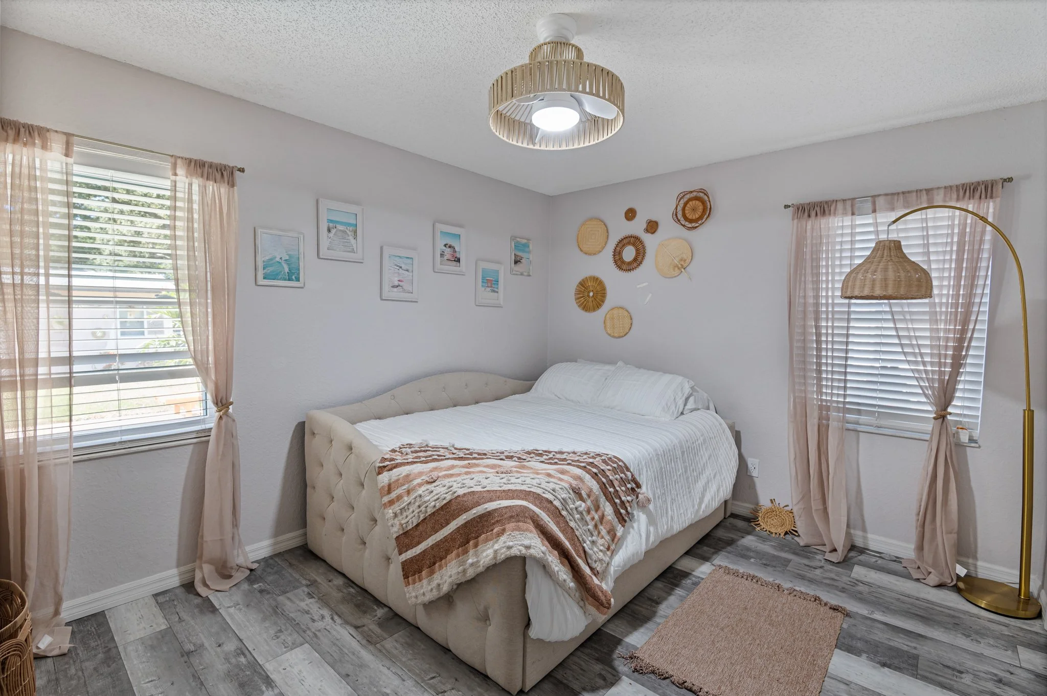 A cozy bedroom with a tufted beige bed, light pink curtains on two windows, and decorative wall art including photos and woven baskets. A wicker lampshade hangs from the ceiling, and a tall floor lamp with a woven shade stands beside one window. The 