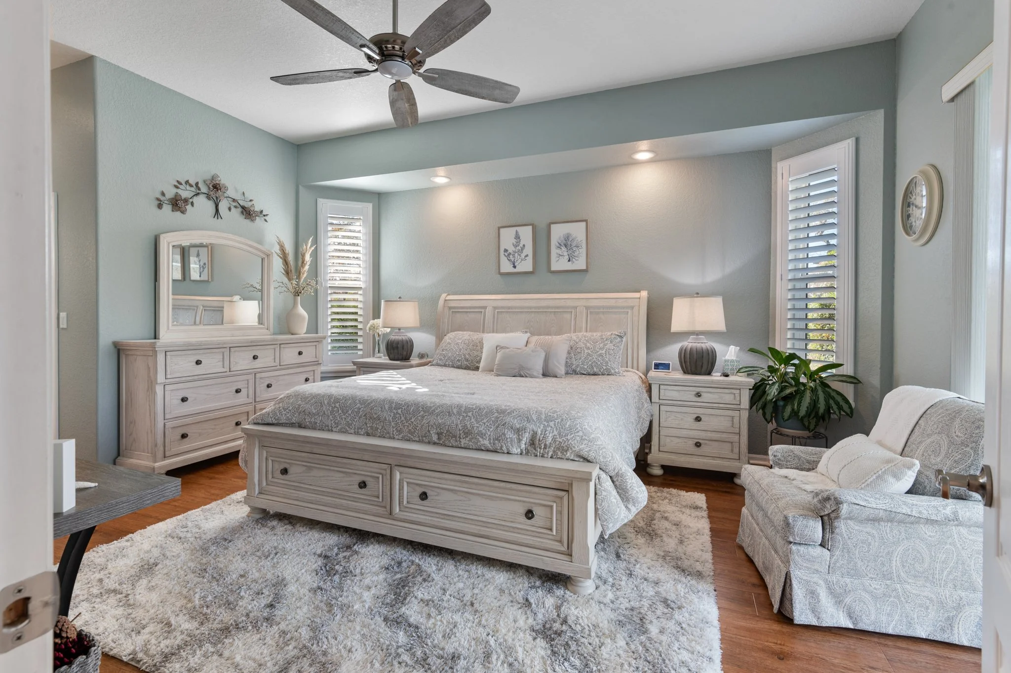 A cozy bedroom with a king-sized bed, two nightstands with lamps, a dresser with a mirror, a small armchair, and decorative wall art and plants, featuring light-colored furniture and blue-green walls.