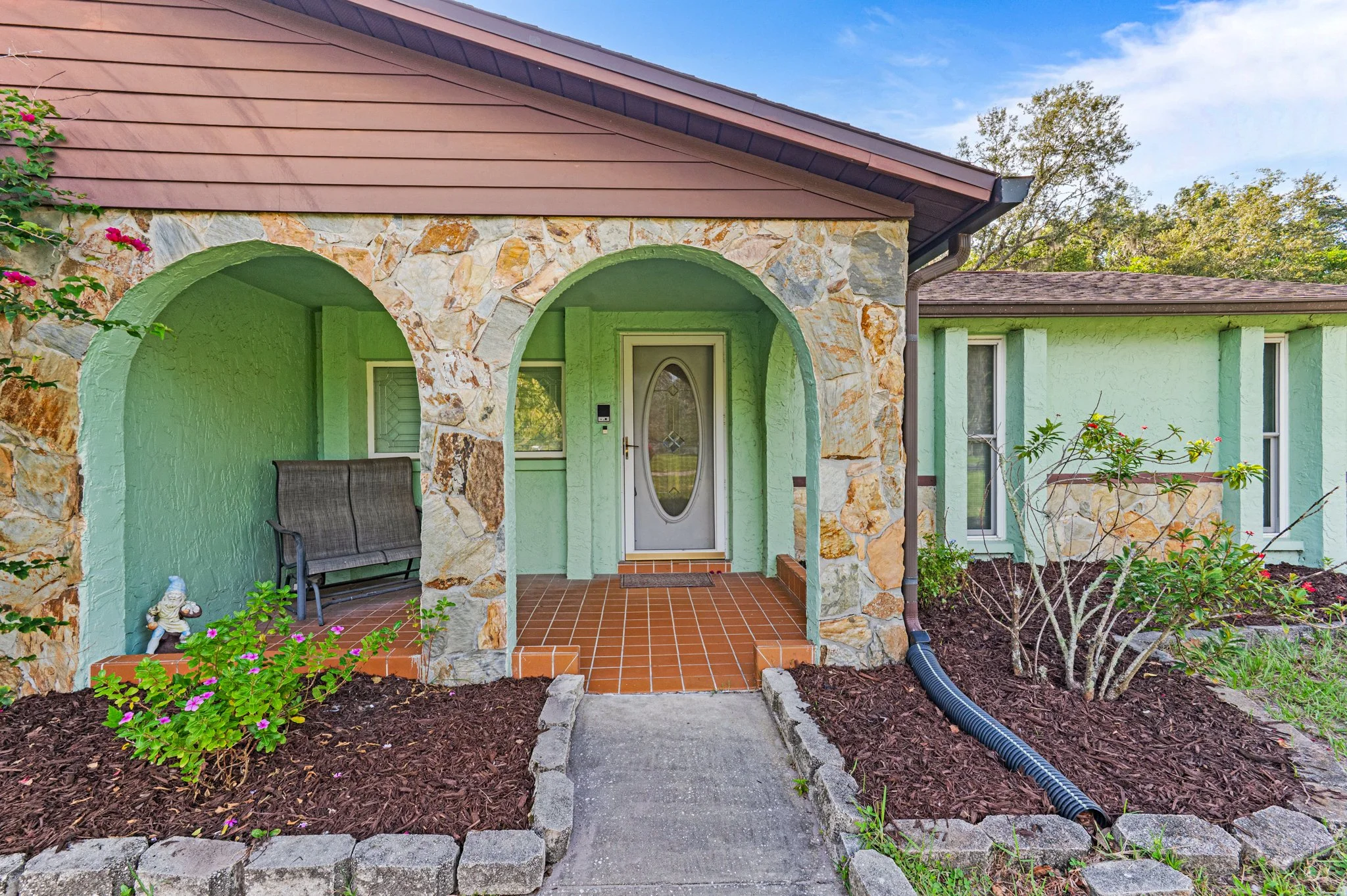 Front porch of a house with arched entryway, stone and green stucco exterior, small garden and bushes, brown tiled porch floor, and a white door.