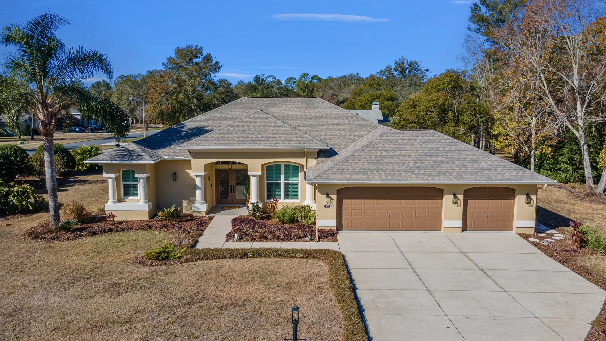 A single-story house with a three-car garage, beige walls, green shutters, and a shingled roof, surrounded by landscaped yard with plants, trees, and a concrete driveway.