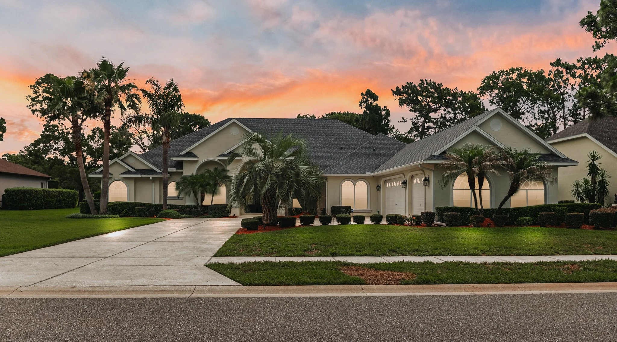 A suburban house with a gray roof, white walls, and large windows, surrounded by palm trees and neatly trimmed bushes, during sunset with a colorful sky.