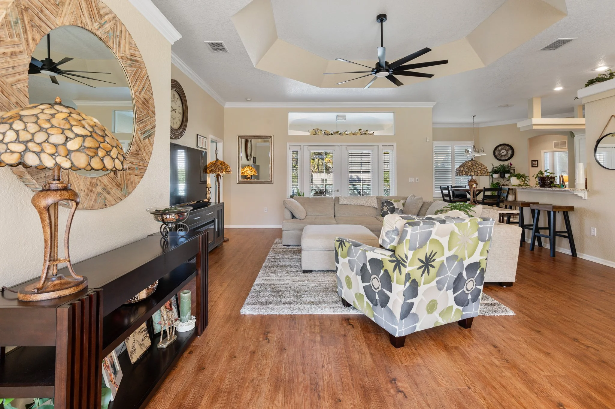 Spacious living room with beige walls, hardwood floors, and ceiling fans. Features a sectional sofa, floral armchair, TV on a black entertainment center, and decorative lamps. French doors with white shutters lead outside, and there is a dining area 
