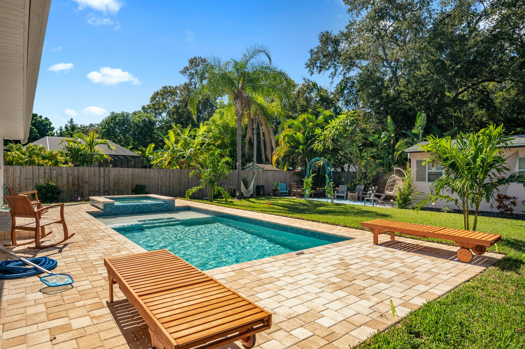 A backyard with a swimming pool, surrounded by a brick patio, green grass, and lush tropical trees. There are wooden lounge chairs, a garden hose, outdoor seating, and a fence with trees and plants.