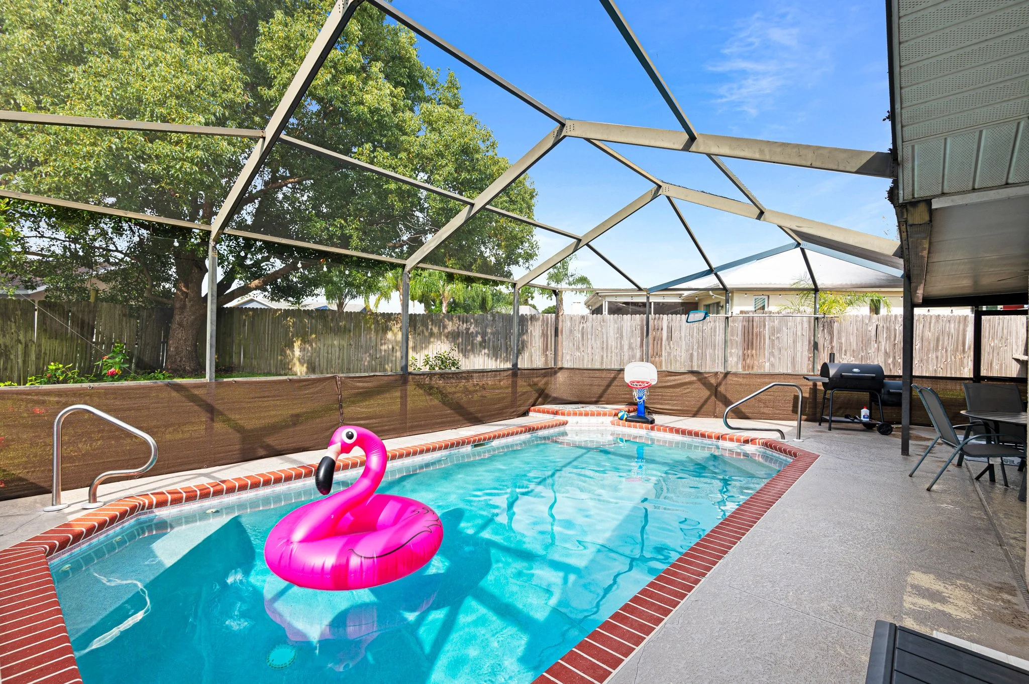 Enclosed backyard swimming pool with a pink flamingo float, a small basketball hoop, barbecue grill, patio chairs, and table, surrounded by a wooden fence and trees, under a clear blue sky.