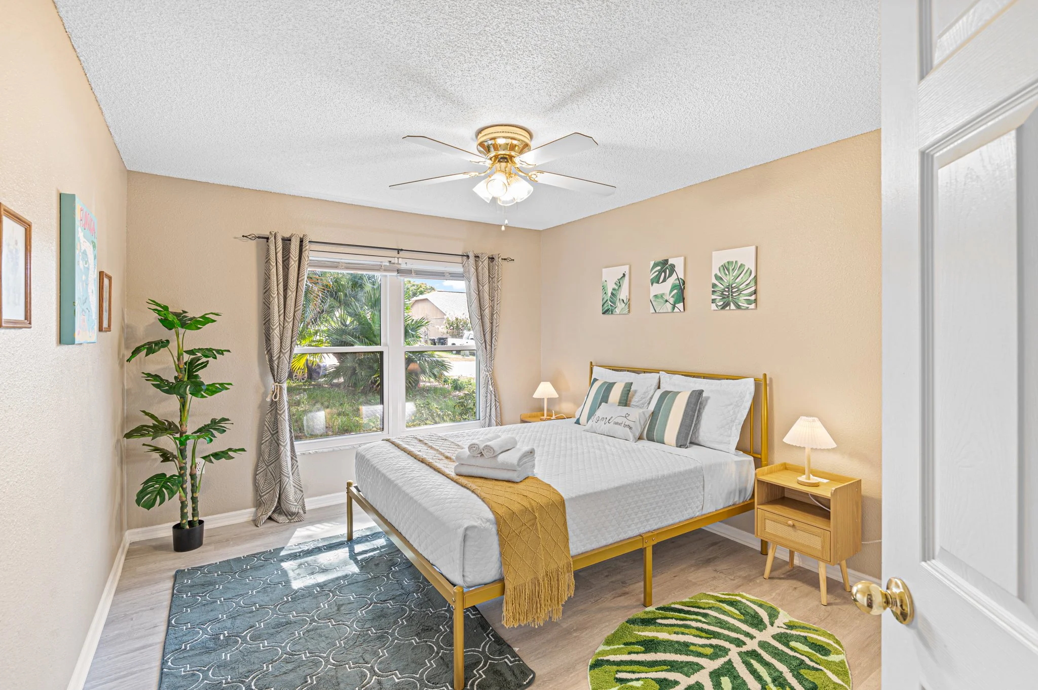 Bright bedroom with beige walls, large window with patterned curtains, queen bed with white bedding and decorative pillows, yellow metal bed frame, wooden nightstands with lamps, indoor plant, wall art featuring tropical leaves, ceiling fan with ligh