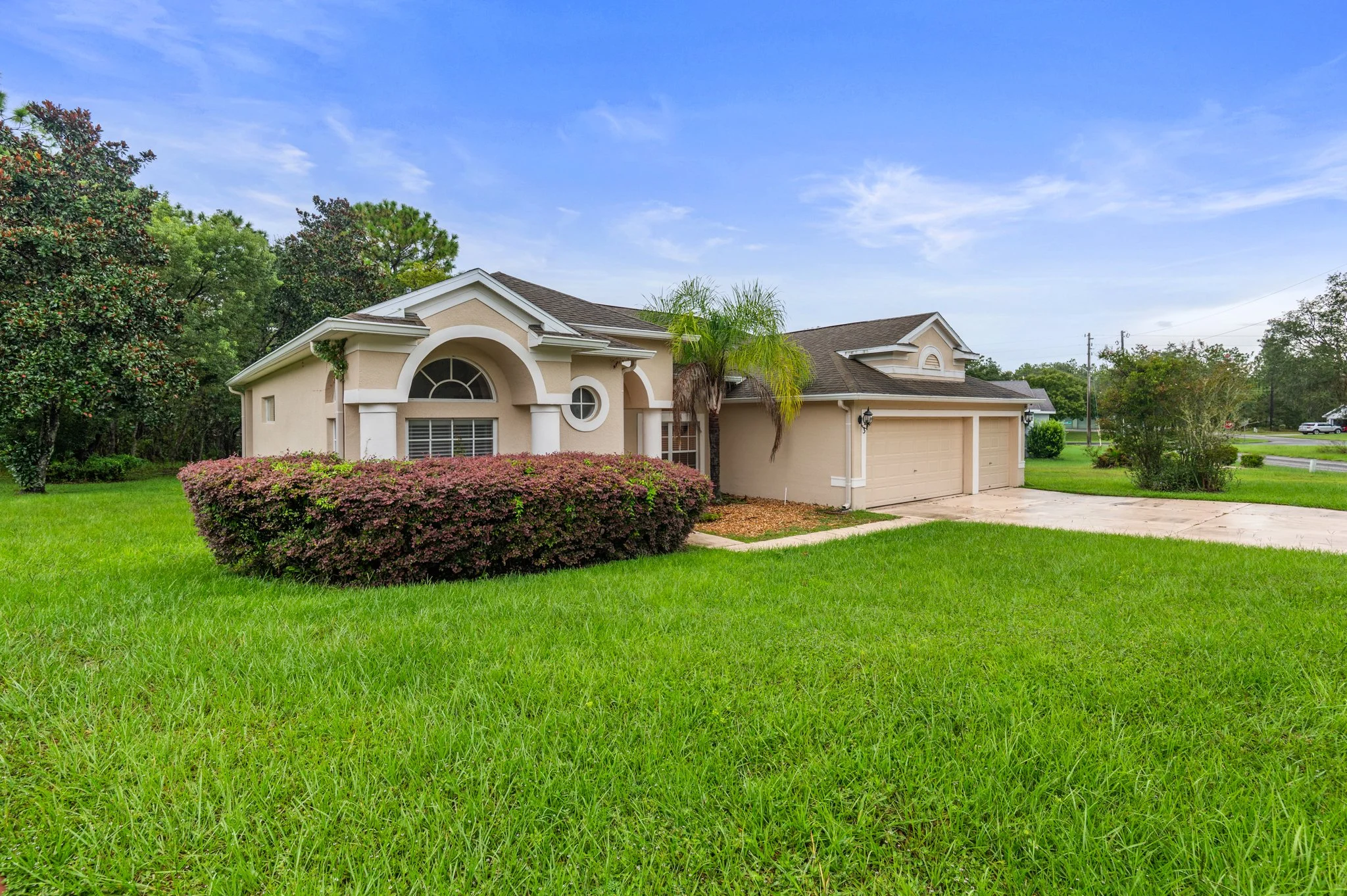 Single-story suburban house with beige exterior, green lawn, bushes, and trees under a partly cloudy sky.