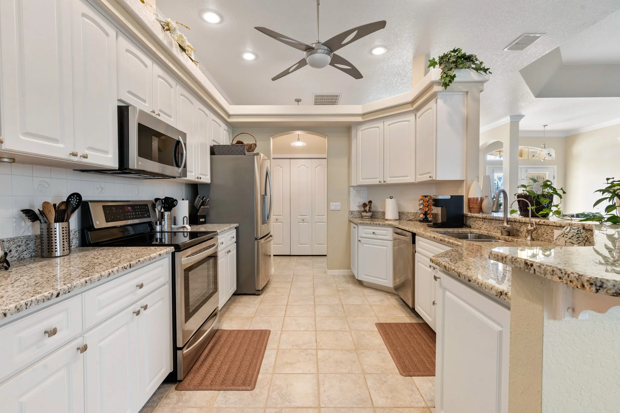 Kitchen with white cabinets, granite countertops, stainless steel appliances, tile floor, ceiling fan, and potted plants.