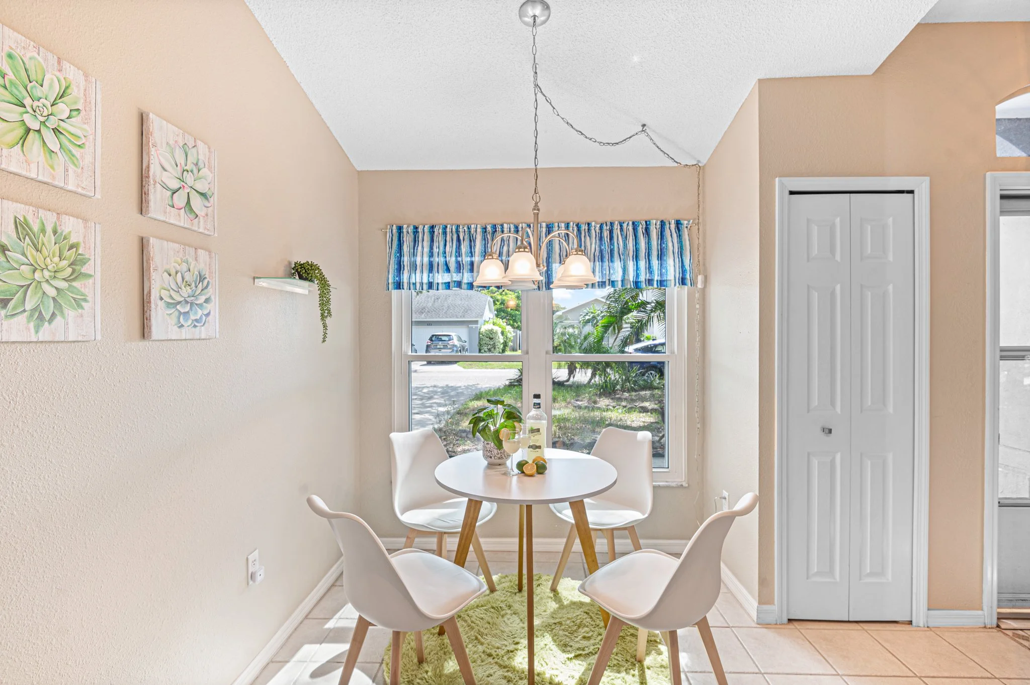 Bright dining area with round white table, four white chairs, a green rug, a window with striped blue curtains, a plant and lemon slices on the table, and wall art of succulent paintings.
