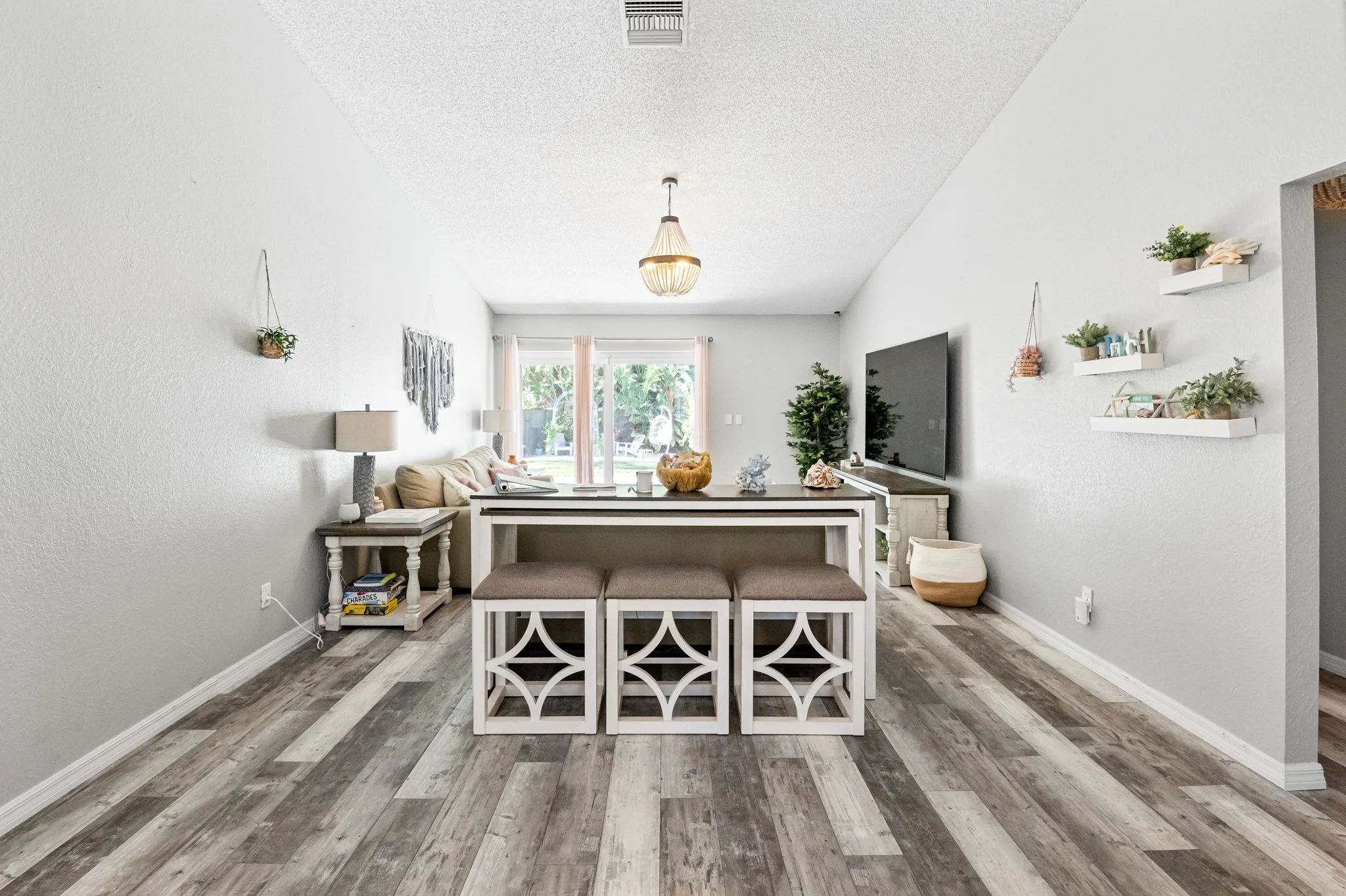 Living room with white walls, hardwood floors, a beige sofa, two side tables with lamps, a flat-screen TV on a white cabinet, a dining table with three stools, and sliding glass doors leading to a backyard with trees.