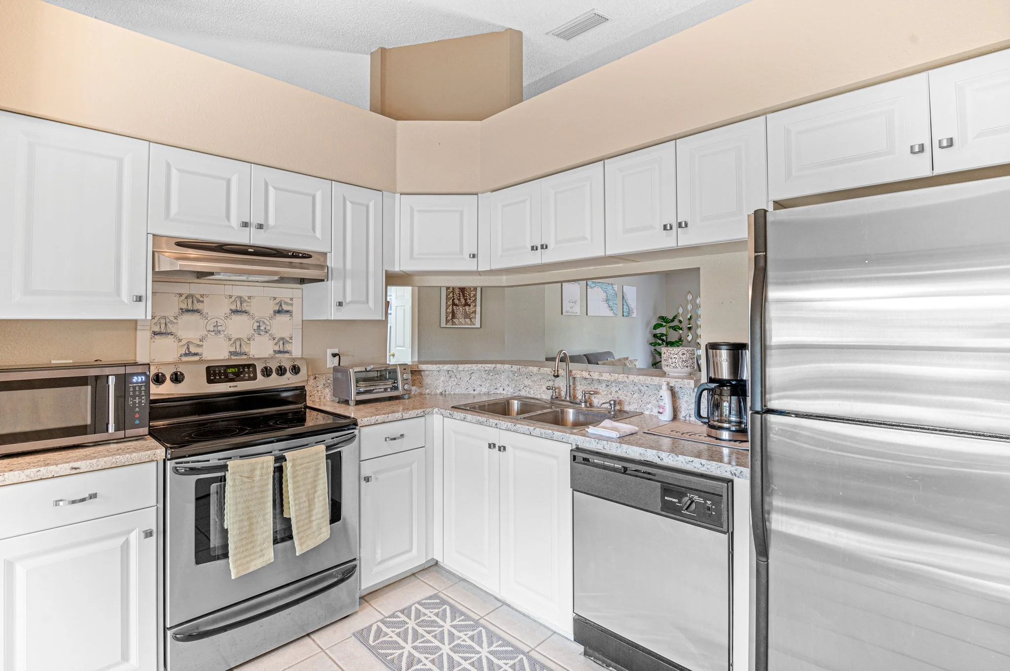 Kitchens with white cabinets, stainless steel appliances including microwave, stove, dishwasher, and refrigerator, a double sink, a small countertop toaster oven, coffee maker, and decorative wall tiles with nautical designs.