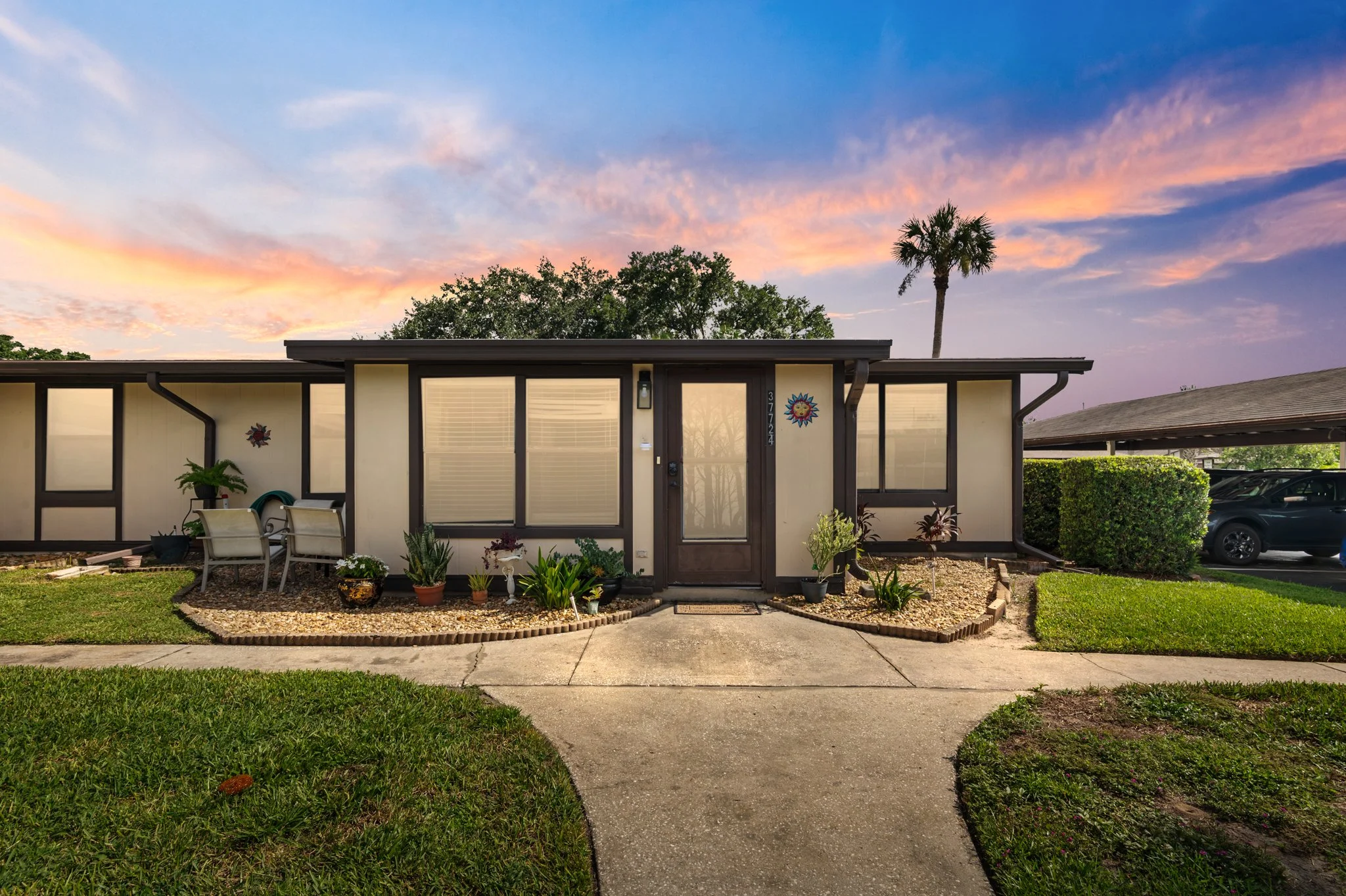 A single-story house with a mid-century modern design, featuring large windows and a slim front door. The front yard has a curved concrete walkway, various potted plants, and a small patio with outdoor chairs. A palm tree and lush greenery are visibl