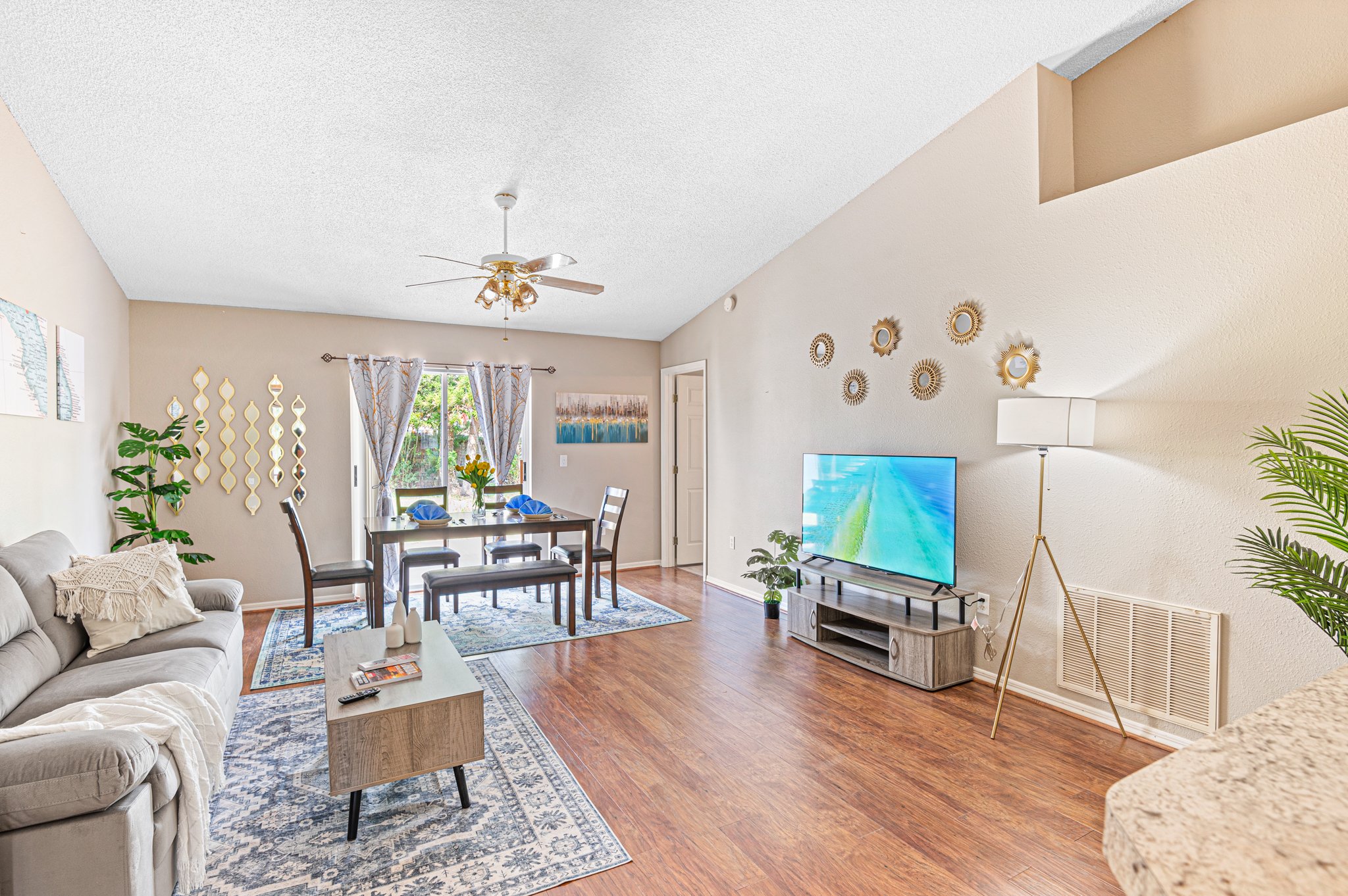 Bright living room with beige sofa, wooden coffee table, TV on wooden stand, dining table with chairs near window, and decorative wall mirrors.