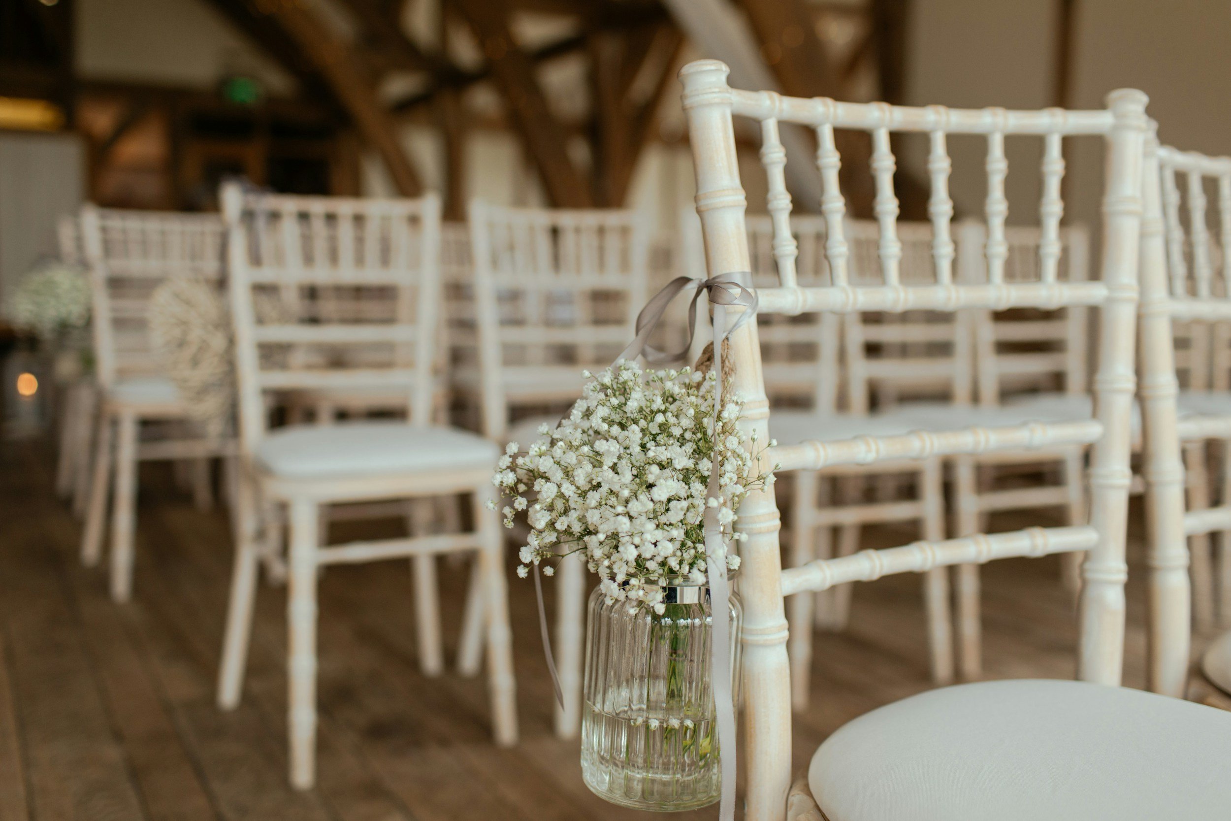 White Chiavari chair with a small vase of white baby's breath flowers hanging from the backrest, in a room with multiple similar chairs arranged in rows.