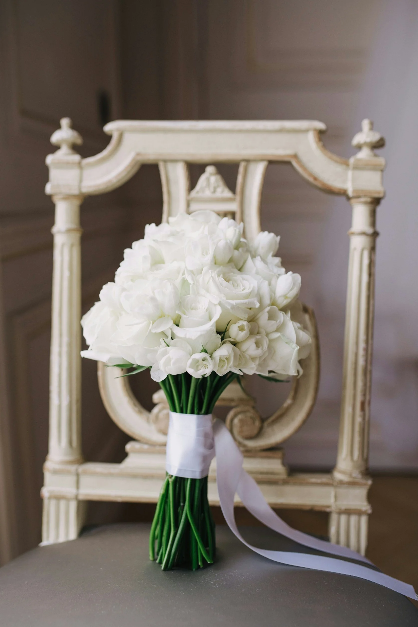 A bouquet of white flowers, including roses and tulips, tied with a white ribbon, placed on a gray surface in front of an ornate wooden chair with a distressed cream finish.
