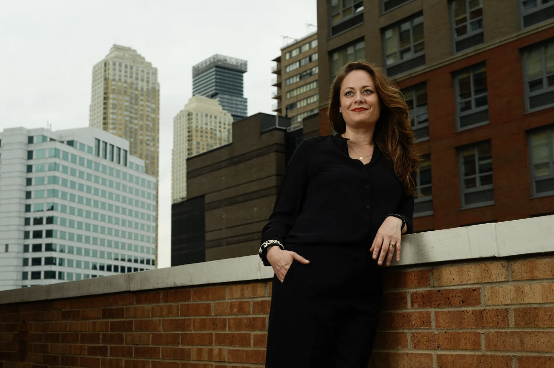 A woman with long hair, wearing a black outfit, standing on a rooftop with city skyscrapers in the background.