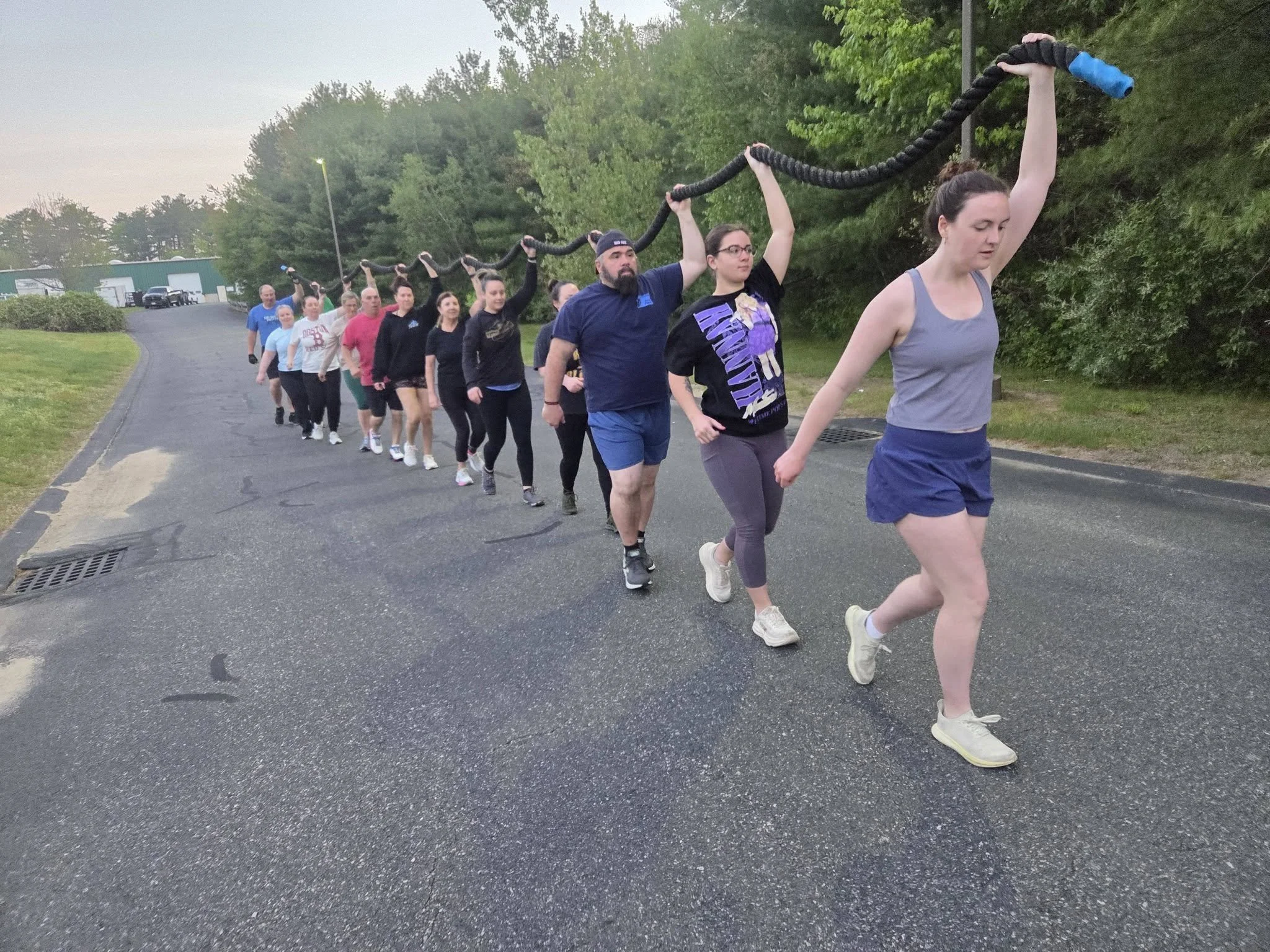 A group of people walking in a line outdoors on a paved path, holding a large rope with black and blue detailing. There are trees and a few buildings in the background.