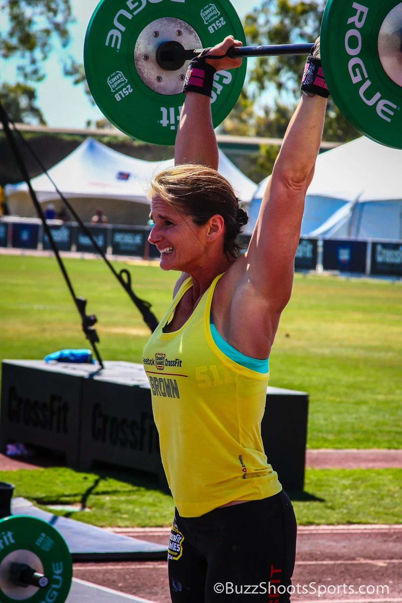 A woman in a yellow CrossFit tank top lifting a barbell overhead at an outdoor CrossFit event, smiling and showing effort.