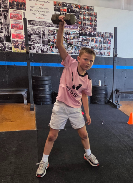 A young boy lifting a dumbbell above his head in a gym, wearing a pink t-shirt and white shorts, with photographs and posters on the wall behind him.