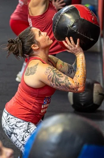 A woman with tattoos wearing a red tank top and patterned leggings is preparing to throw a medicine ball during a workout in a gym.