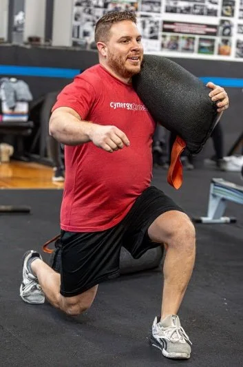 A man in a red shirt and black shorts kneels on one knee in a gym, holding a black foam roller. He appears to be exercising or stretching.