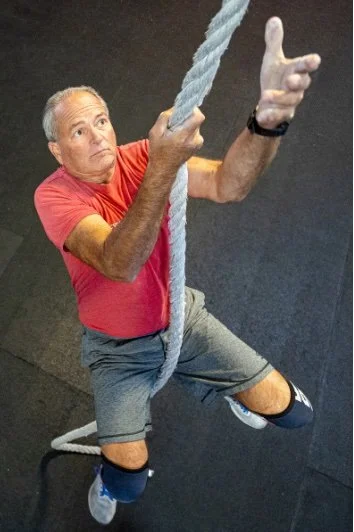 A man in a red shirt and gray shorts climbing a thick rope at a gym, wearing knee braces and sneakers, with a focused expression.