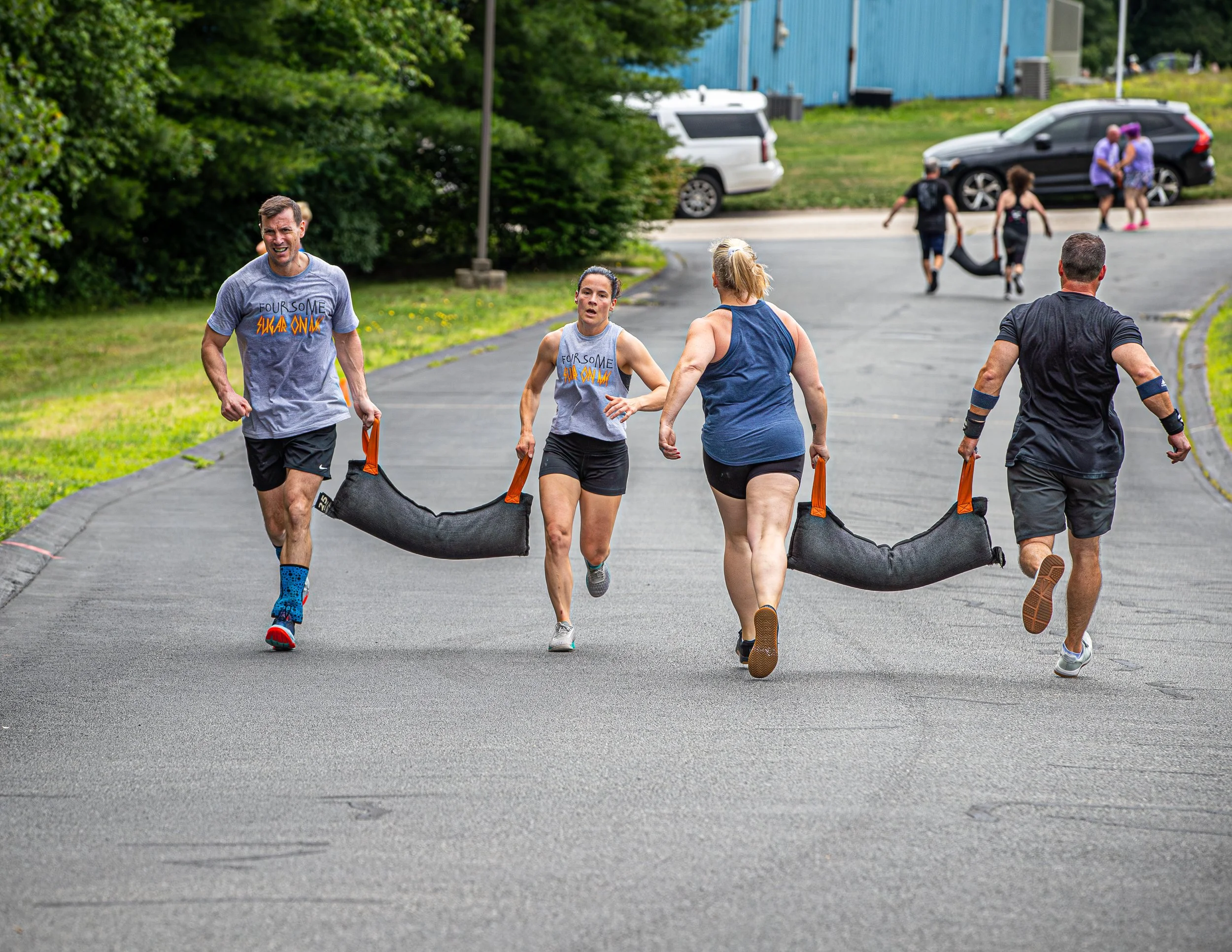 Group of runners participating in a race or event on an outdoor paved path, surrounded by greenery and parked cars.
