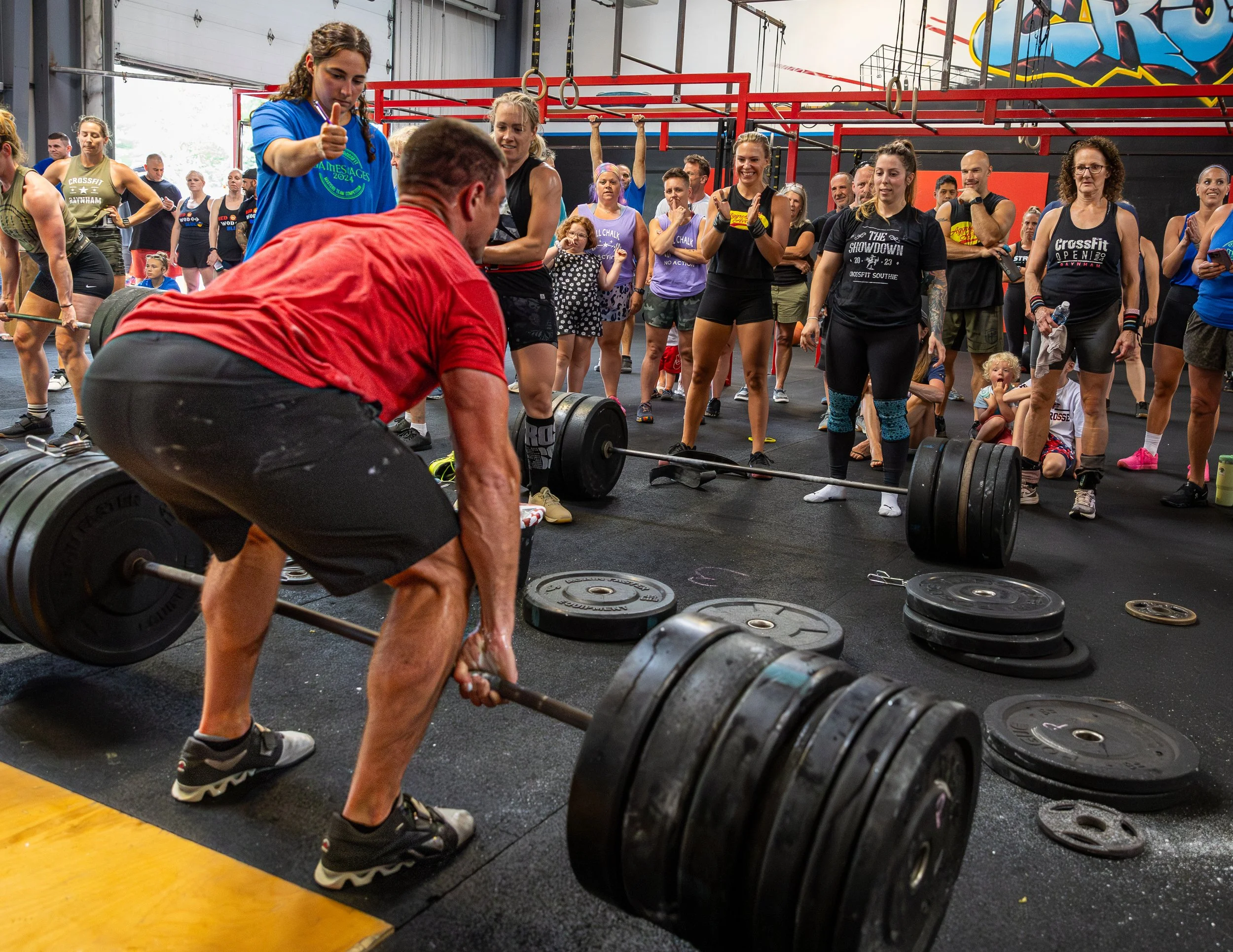 A woman in a red shirt and black shorts lifts a barbell with weights while a crowd of people watches at a CrossFit gym.