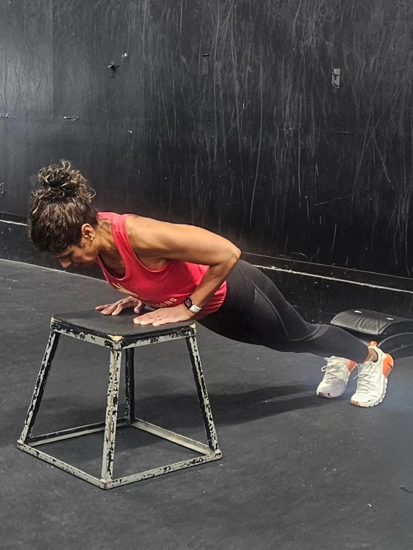 Woman in a red tank top and black leggings doing push-ups on a black floor, using a worn-out black step stool for support in a gym or fitness studio.