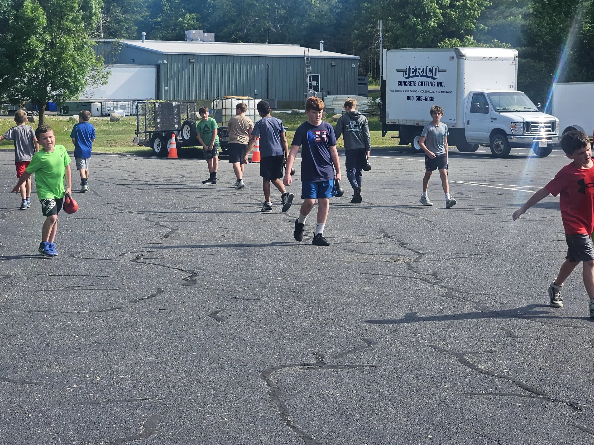 A group of young boys walking and playing in a parking lot on a sunny day, with a blue industrial building, a white truck with business information, and green trees in the background.  Youth strength and conditioning, agility and speed training