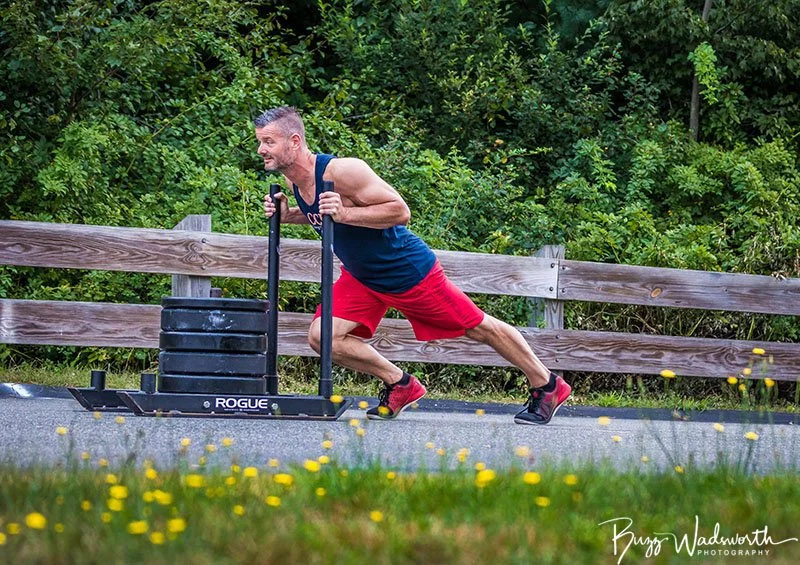 A man in a blue tank top and red shorts pushes a weighted sled on a paved path surrounded by green trees and a wooden fence, with yellow flowers in the grass nearby.