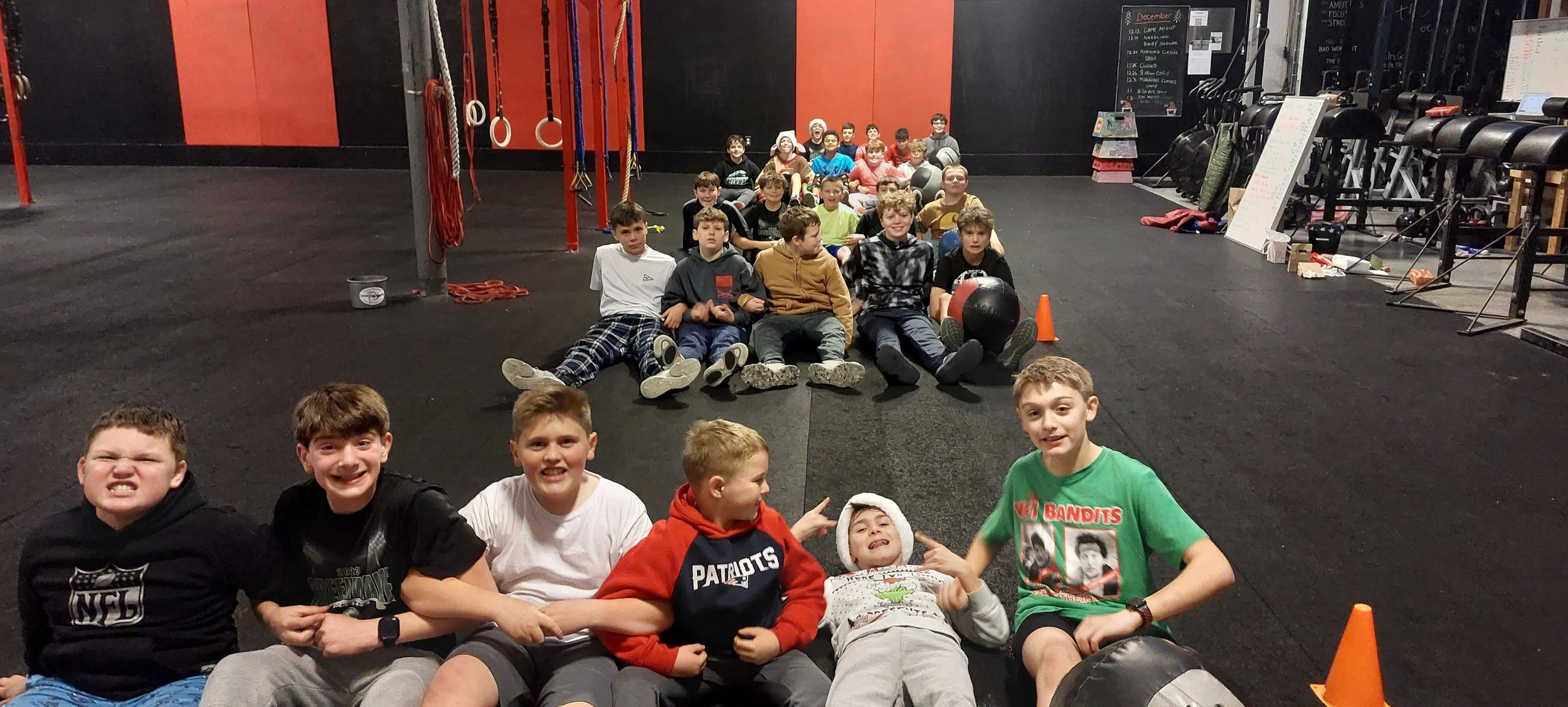 A group of boys sitting on the floor in a gym, with some lying down and others sitting, and a large group of kids in the background sitting in a line, inside a gym with fitness equipment and black and red walls.